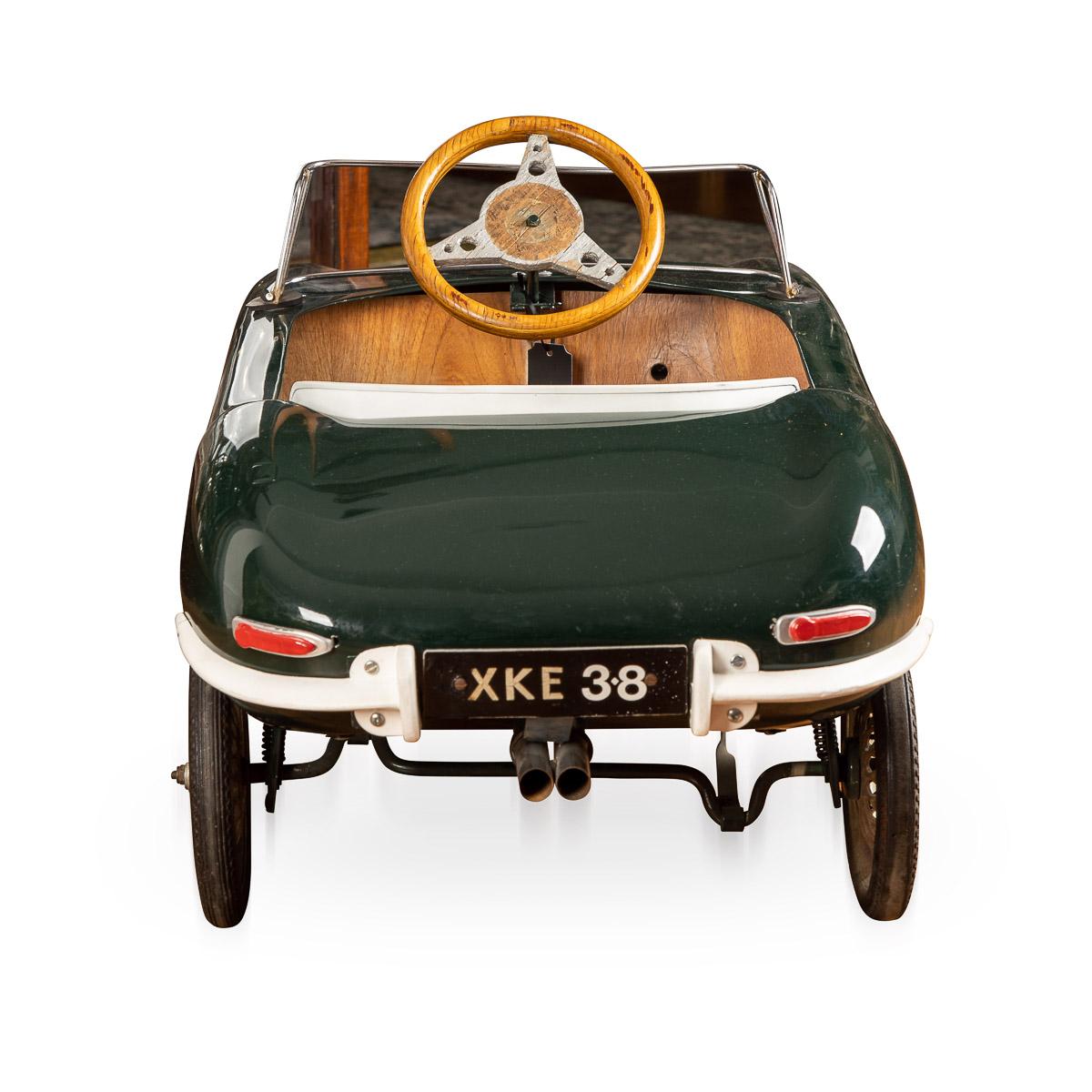 A mid-20th century English made “Jaguar E-Type“ model pedal car, green steel body on metal framework with white metal panel seat, treadle pedals to cranked rear axle, wooden steering wheel, number plate to rear bumper reads XKE 3.8, cast alloy wheel