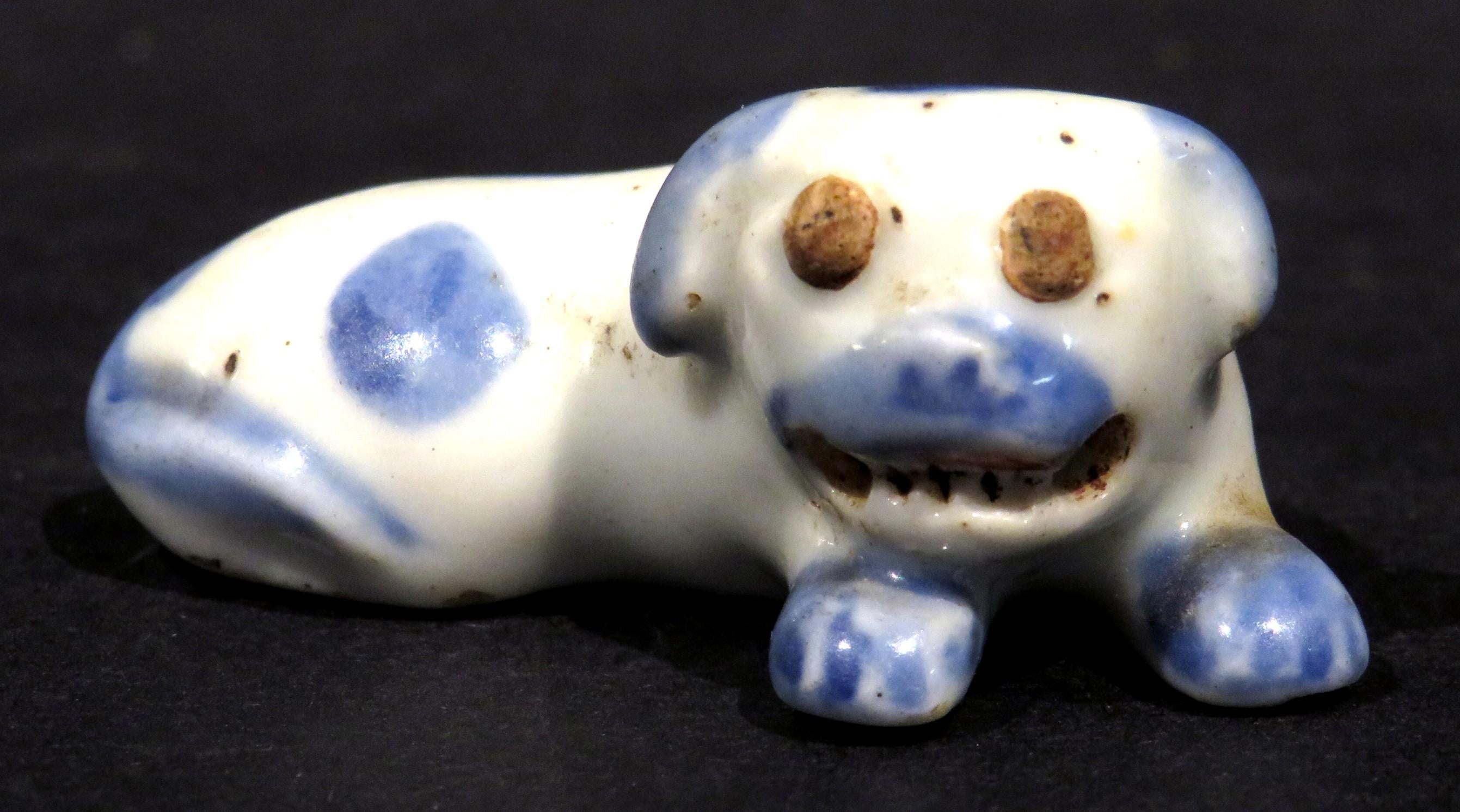 The recumbent white porcelain figure of a pug dog shown baring its teeth, decorated by hand with spot painted cobalt detail, the underside with twin holes intended for a cord to pass through & attach it to an obi (sash). Measuring, .7/8