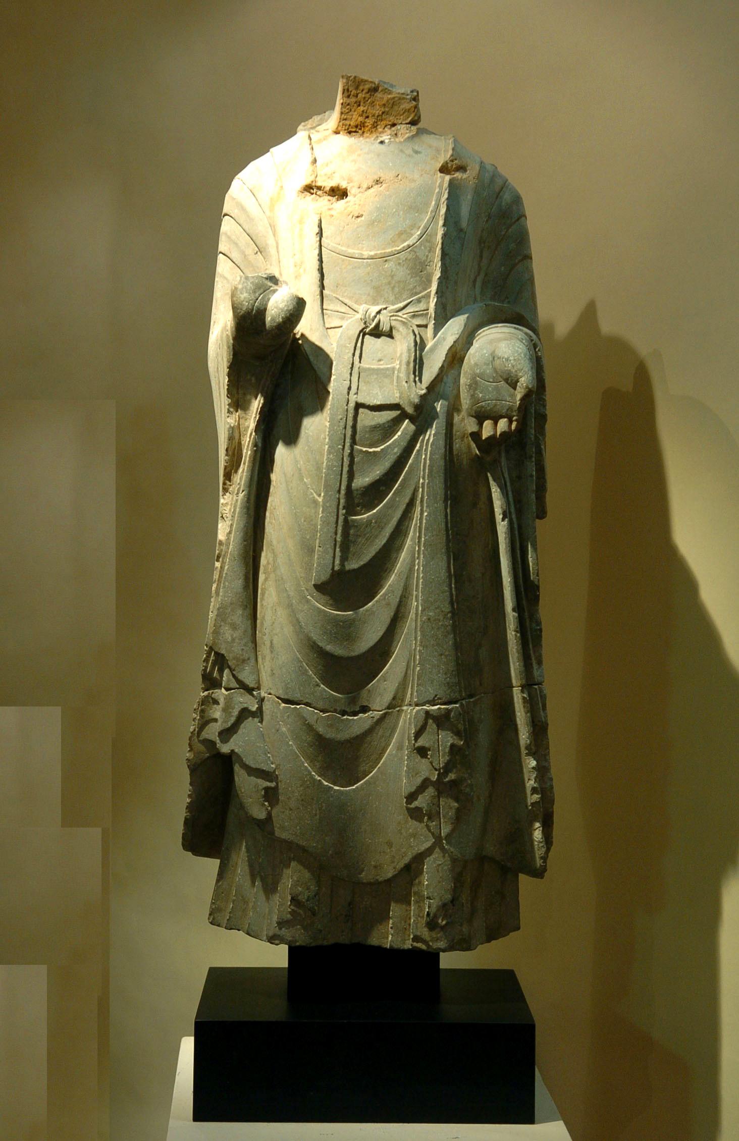 EINE MASSIVE BUDDHA-FIGUR AUS KALKSTEIN 
Die imposante stehende Figur trägt ein einfaches Gewand, das die Konturen des Körpers subtil erkennen lässt, mit U-förmigen Falten und anmutig über den linken Arm drapiert, der Rücken ist bis auf ein Stück