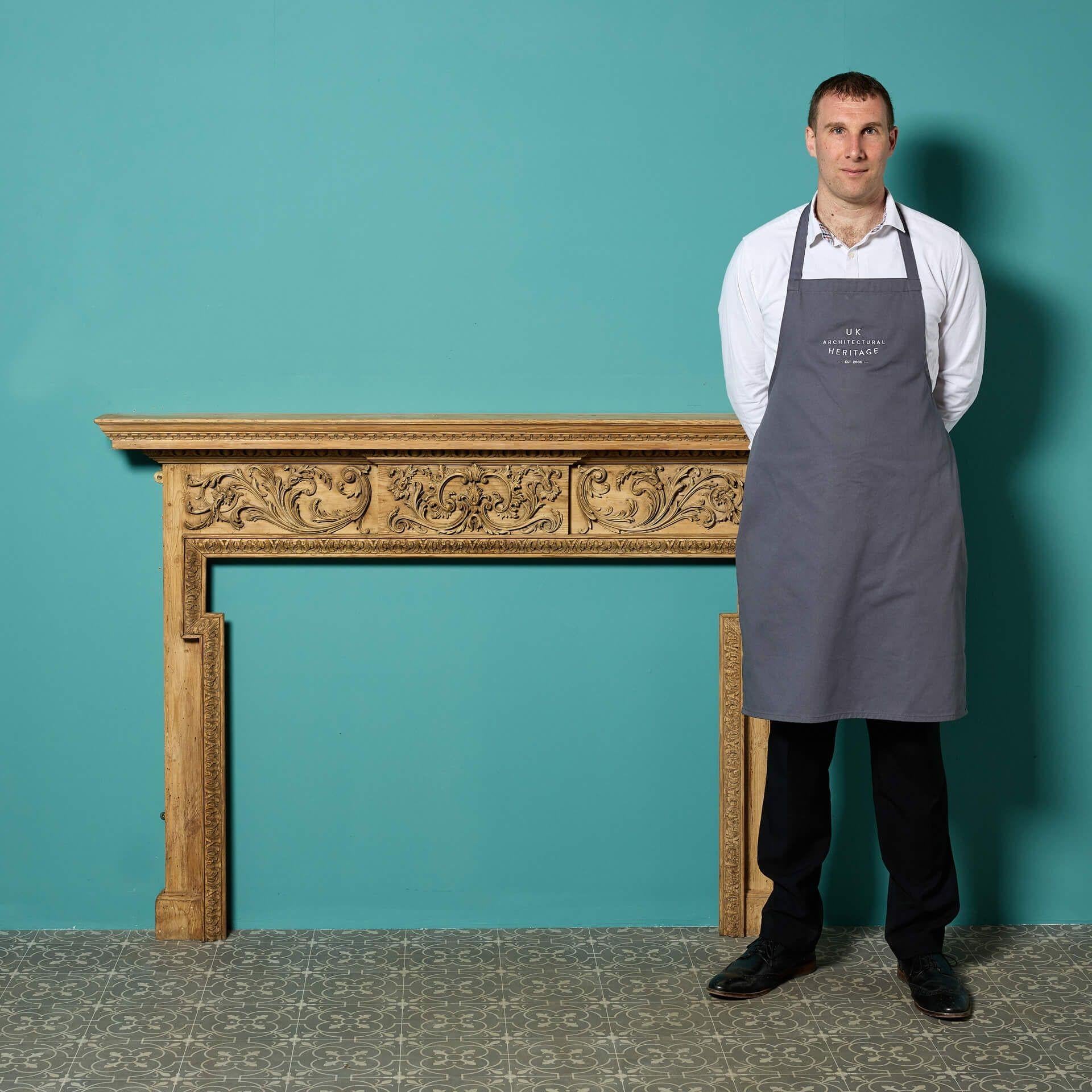 A reclaimed Georgian style carved pine fireplace. Dating to the late 19th century, this fire surround showcases ornate scrolling foliage to the frieze with slender jambs and a unique dog leg opening.

The stripped finish of this fireplace gives it a
