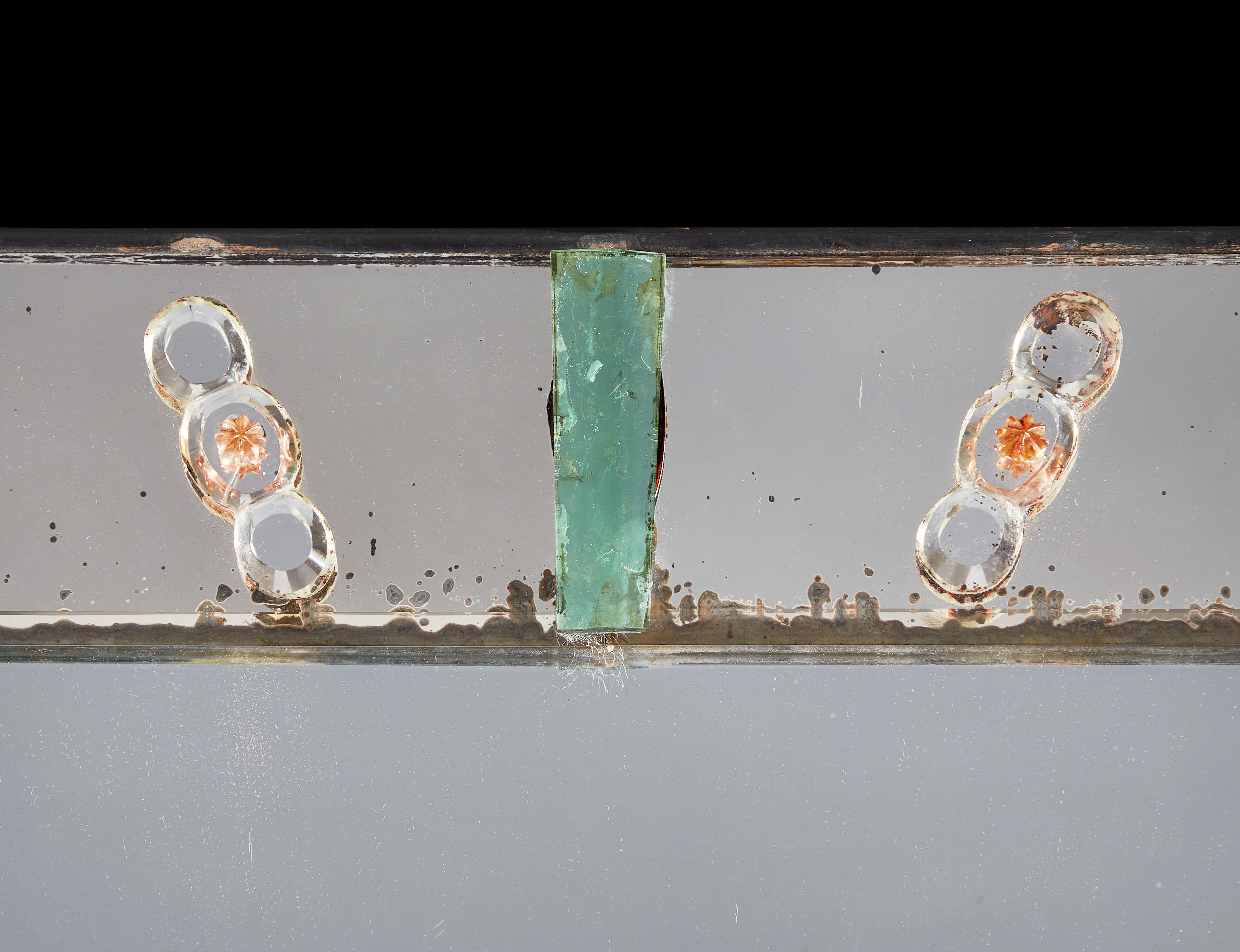 A large rectangular Venetian mirror with beveled frame, decorated with a mixture of glass leaves and two pale green glass pieces at the centre of the top and base. There is age around the edges of the frame but the central glass is clear and gives