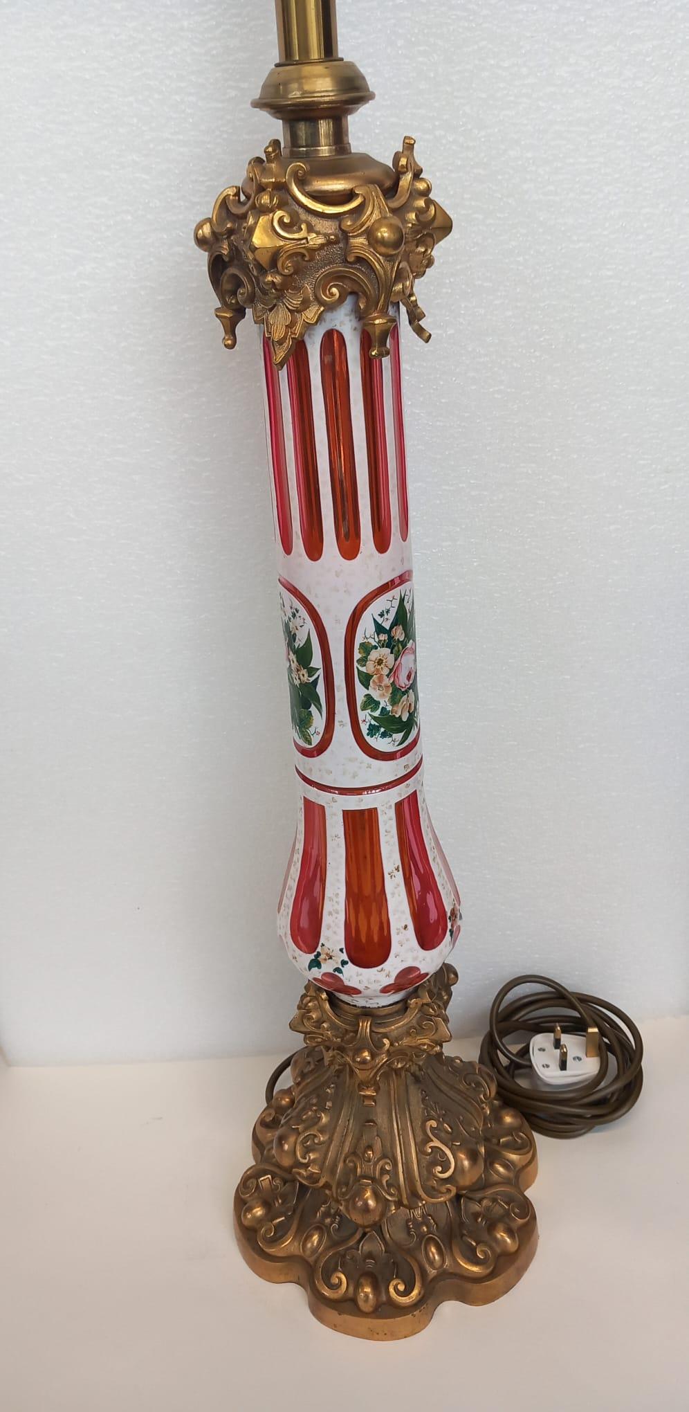 A red Bohemian glass lamp dating from the mid nineteenth century, decorated with a white striped enamel design, hand painted with elaborate flower decoration.
The glass lamp sits on an ormolu gothic style foot or base. 
The top part of the lamp has