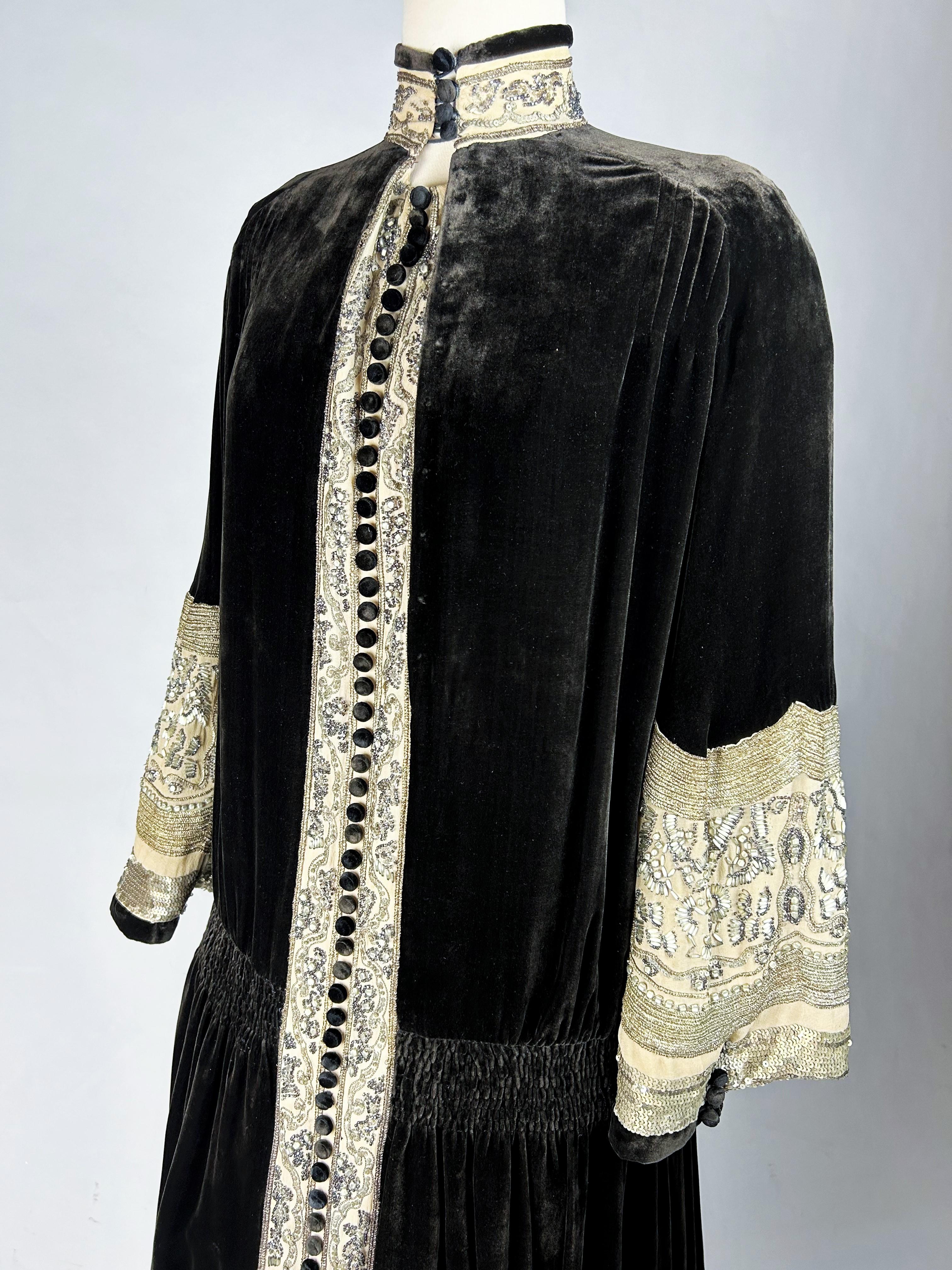 Circa 1923/1924
Paris
Redingote evening dress in chimney sweep brown velvet embroidered in a champagne color by the famous Maison Worth. Ample straight cut, long sleeves, high collar buttoned Russian style, finely gathered in three rows at the