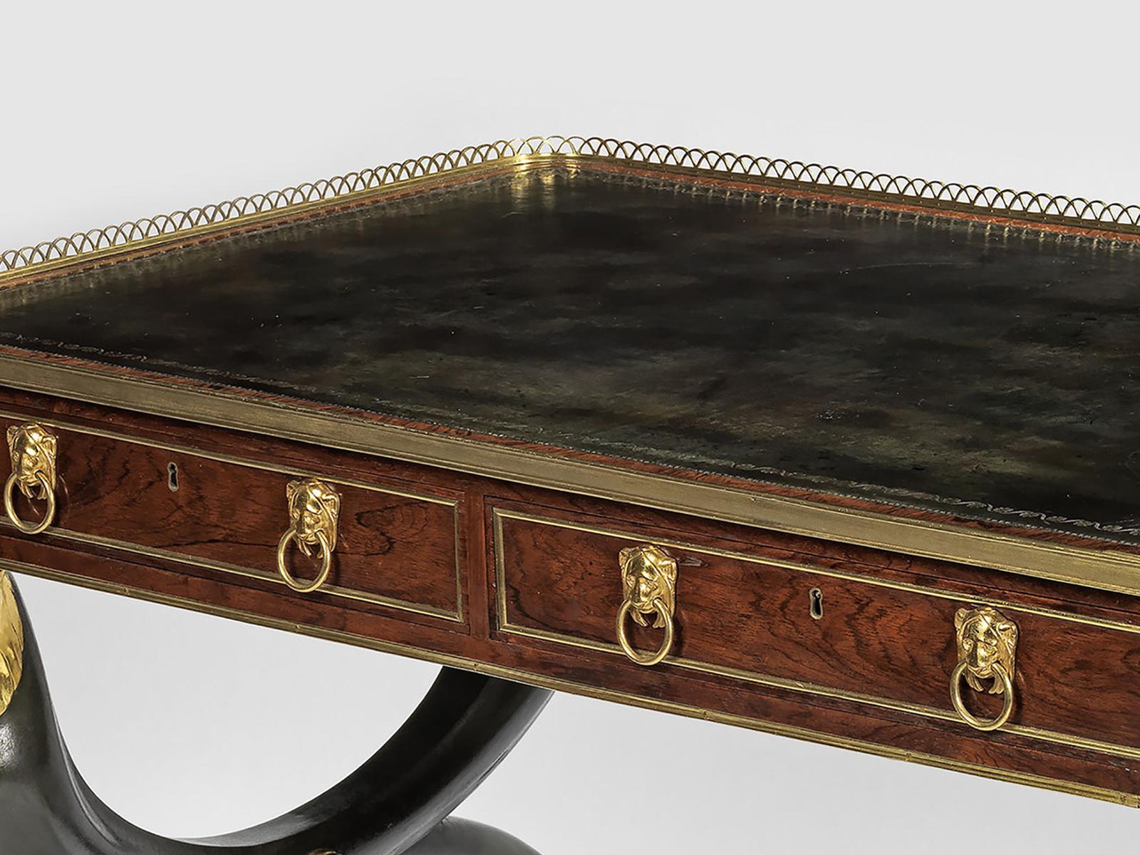 The table has a rectangular top with rounded corners, with a blue leather writing surface surrounded on three sides with a pierced brass gallery. The frieze contains two drawers with lion’s mask drop-ring handles with brass banding; the reverse has