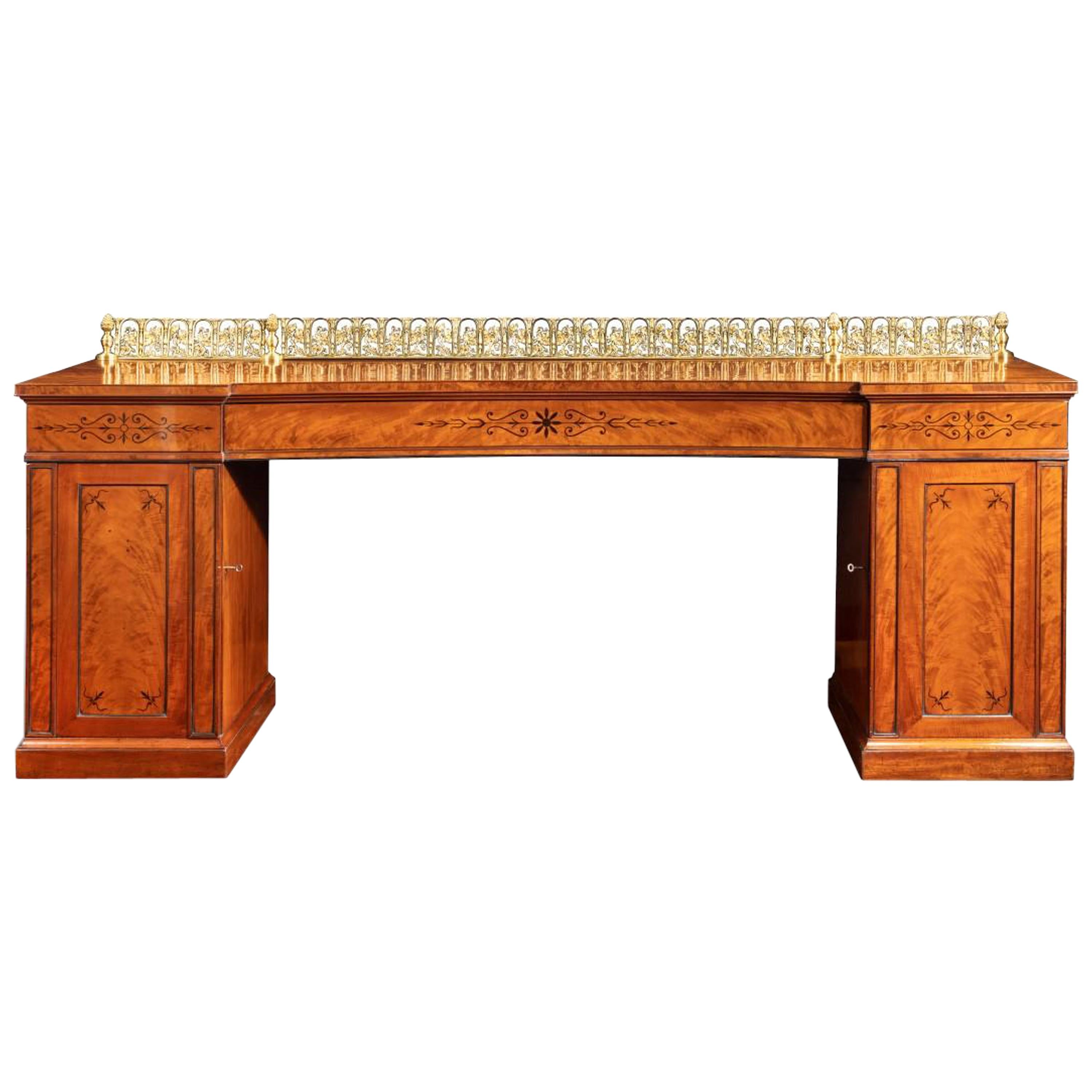 A Regency pale mahogany pedestal sideboard, the rectangular top with a central drawer disguised in the concave frieze and surmounted by a tall ormolu guilloche of arches enclosing vine stems, each pedestal with a cupboard door opening to reveal a