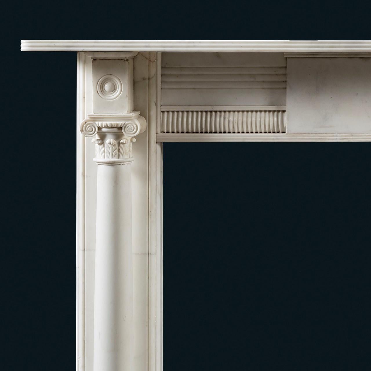 A Regency period statuary marble column chimneypiece, the frieze and shelf enriched with reeds, centred with a plain central tablet, flanked by square corner blocks carved with bullseye roundels, above tapering columns capped by composite capitals,