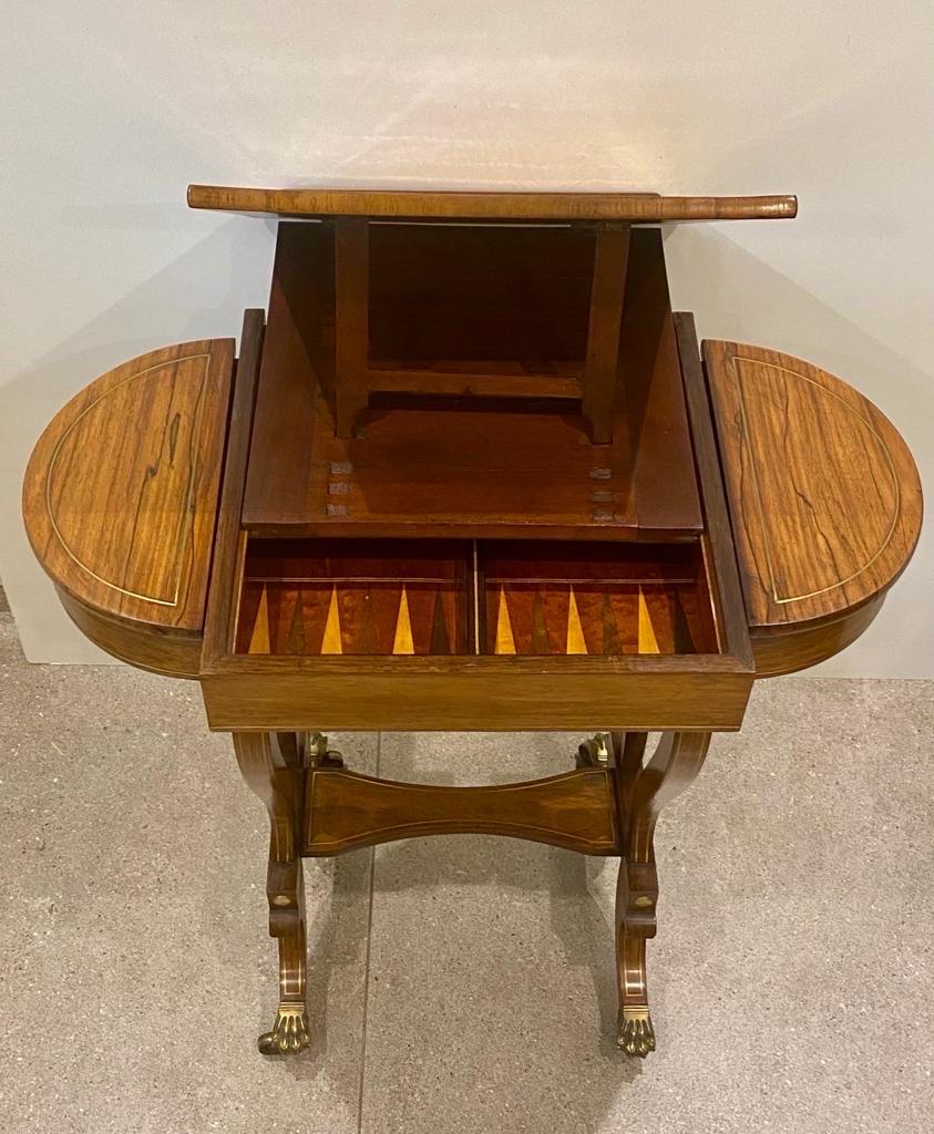 Um 1820, Regency-Spieltisch aus Rosenholz mit Messingbeschlägen und Intarsien. Der Mittelteil mit umkehrbarem Spieldeckel gibt den Blick frei auf ein ausgestattetes Innenleben. Es hat eine zentrale Schublade mit blattförmigen Messingeinlagen.
Dies