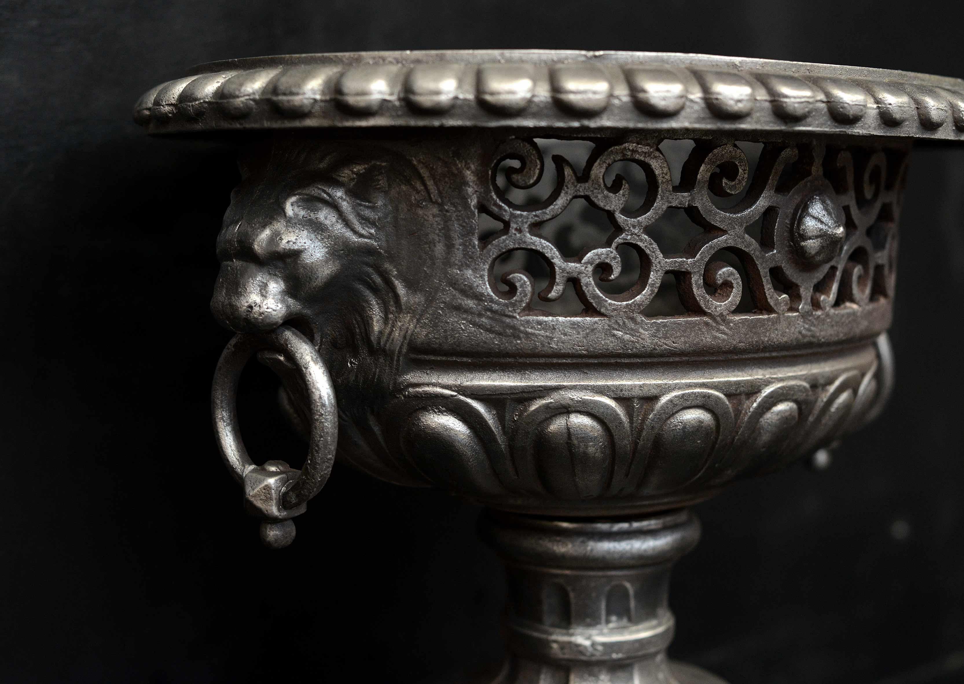 A Regency style urn firegrate in polished cast iron. The gadrooned lower part surmounted by scrollwork and rosette paterae and gadrooned top. The ring handles in lion's masks. Italian Bardiglio marble plinth below. English, 19th