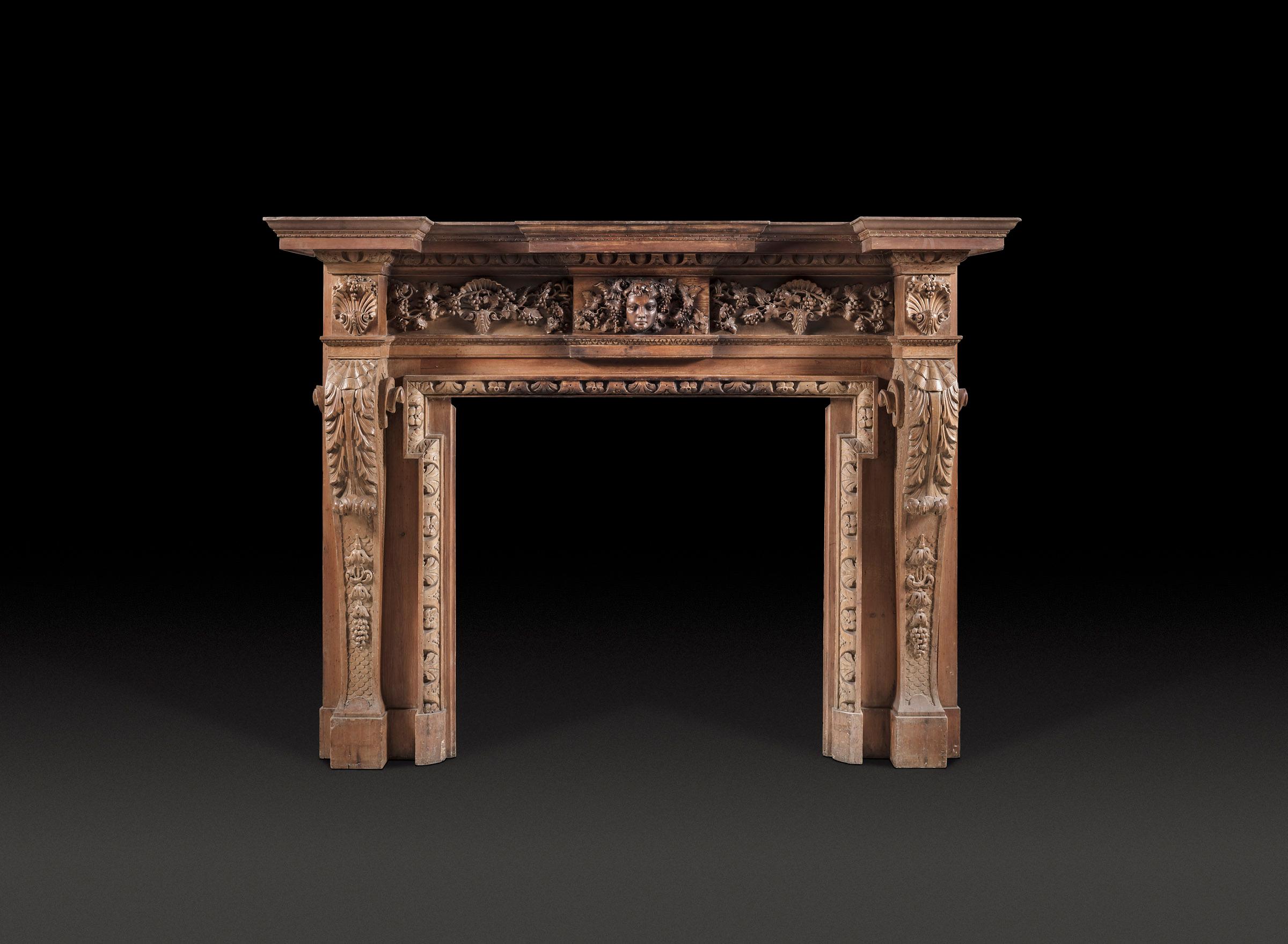 Camino in legno di pino in stile George II del XIX secolo, intagliato in modo robusto e di dimensioni impressionanti alla maniera di William Kent. La tavoletta centrale reca la maschera di Bacco, affiancata da un fregio di viti. I profondi stipiti