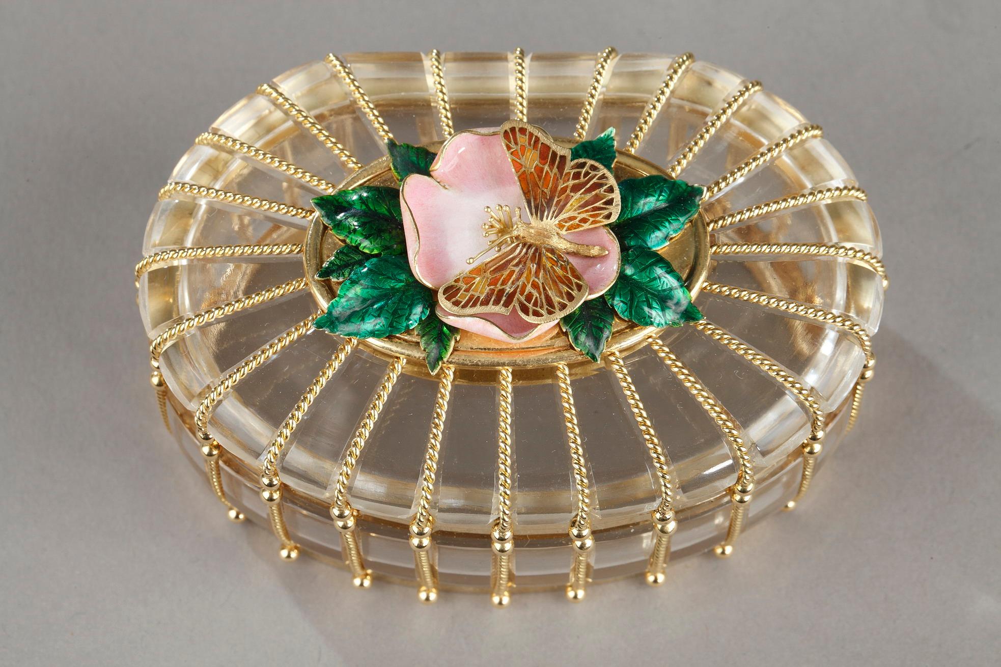 De forme ovale, le couvercle est orné d'une fleur en émail rose et de feuilles vertes sur lesquelles repose un papillon en or aux ailes percées, les montures en cage de corde torsadée, le bord de la base est gravé 
