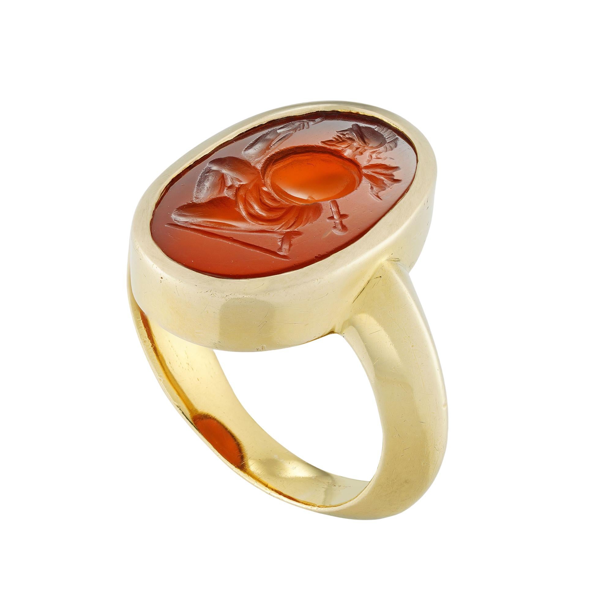 A Roman carnelian intaglio set in a yellow gold ring, the oval-shaped carnelian measuring approximately 18 x 12mm, depicting a kneeling ancient Greek warrior, wearing a helmet with plume, holding a spear and round shield, carved in 1st century BC -