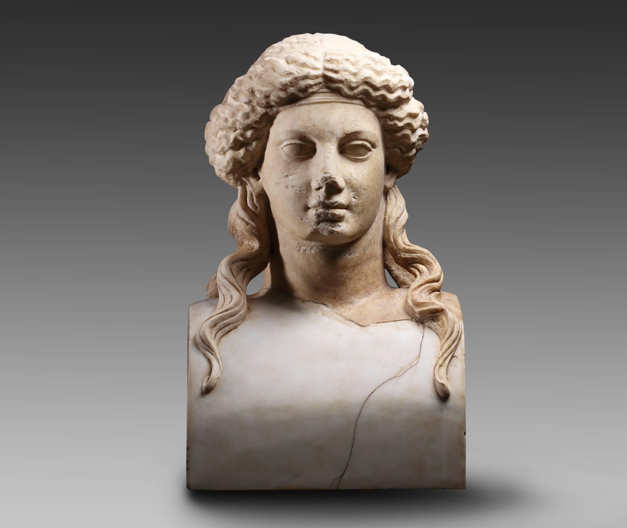 It is a Janiform Roman marble. It consists of two lifesize heads, substantially carved with idealized features.
The cheeks are rounded, the heads have large almond shaped eyes, the wavy hair is swept from their faces and tied in a net. Another net