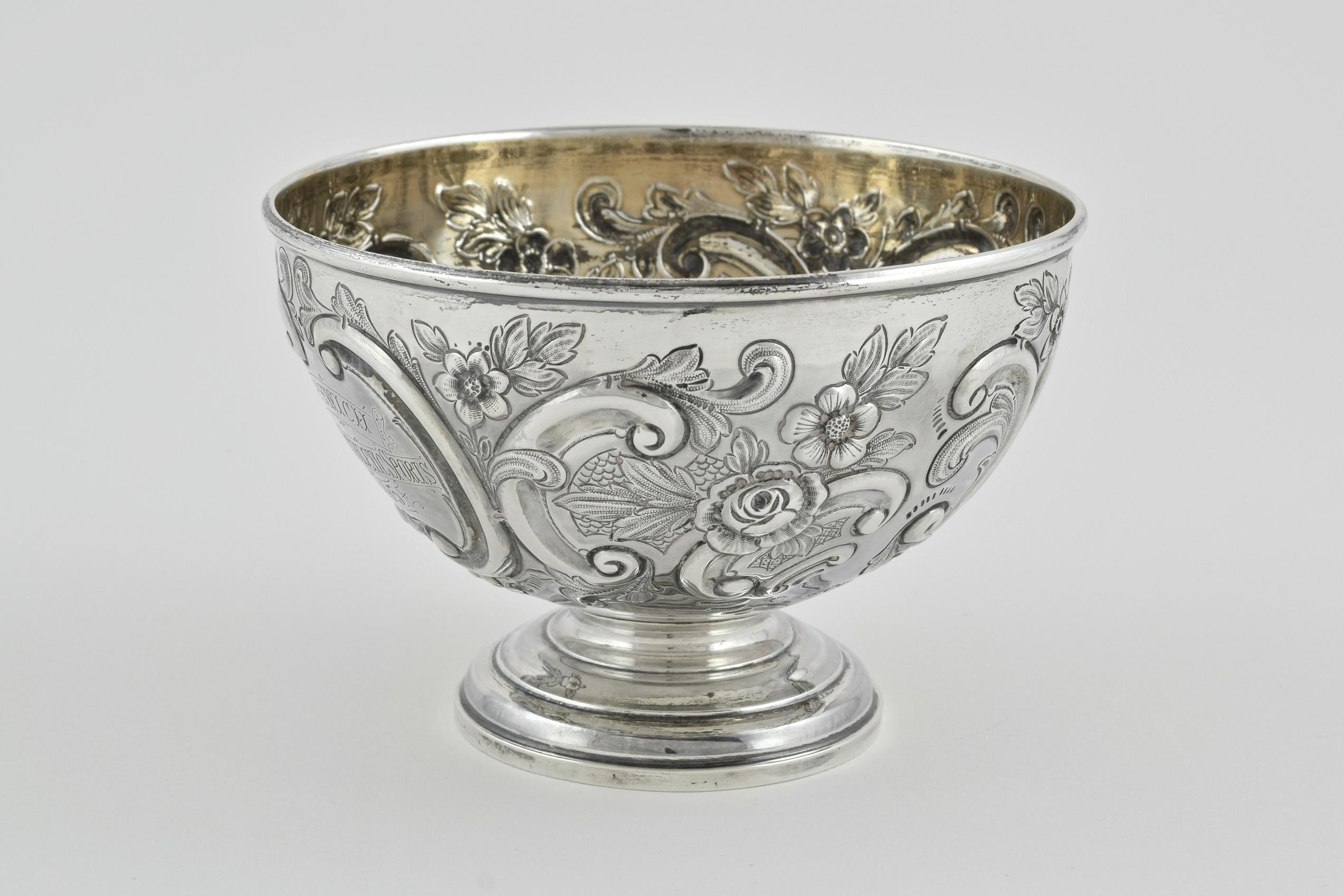 Styled with repousse and chased work, the body of the bowl is Intricately engraved with flowers, foliage and scrolls. The engraving on the body of the bowl reads 'IP Switch Amateur Athletic Sports 1895', and the other 'Two Miles Flat Race won By G.W