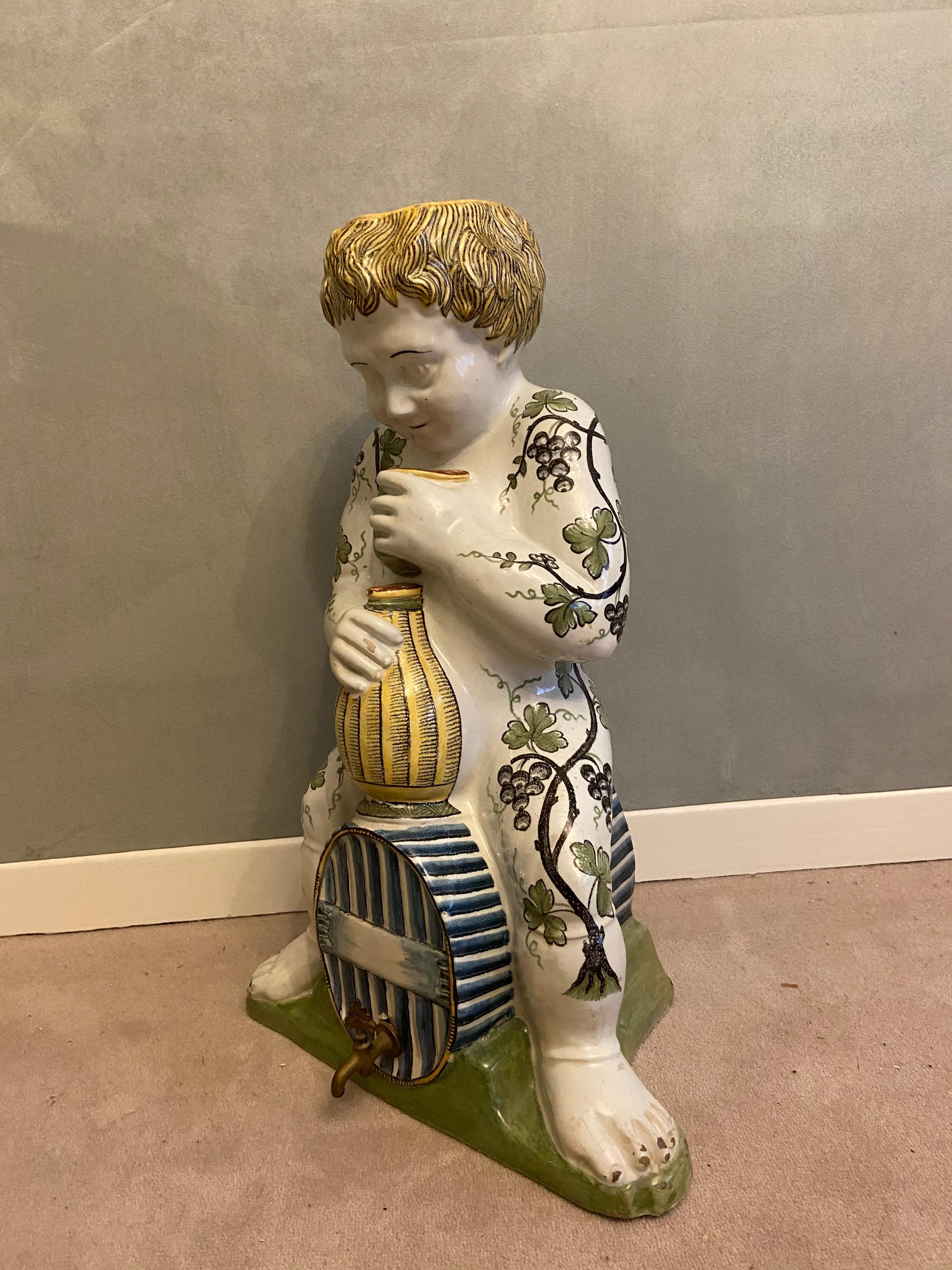 A Rouen Faience in the shape of a young bacchus seated on a blue barrel holding a bottle in his right hand and a glass in his left hand. Polychrome decoration of vine branches and grapes. Rouen Faience. French Work. Circa 18th Century.