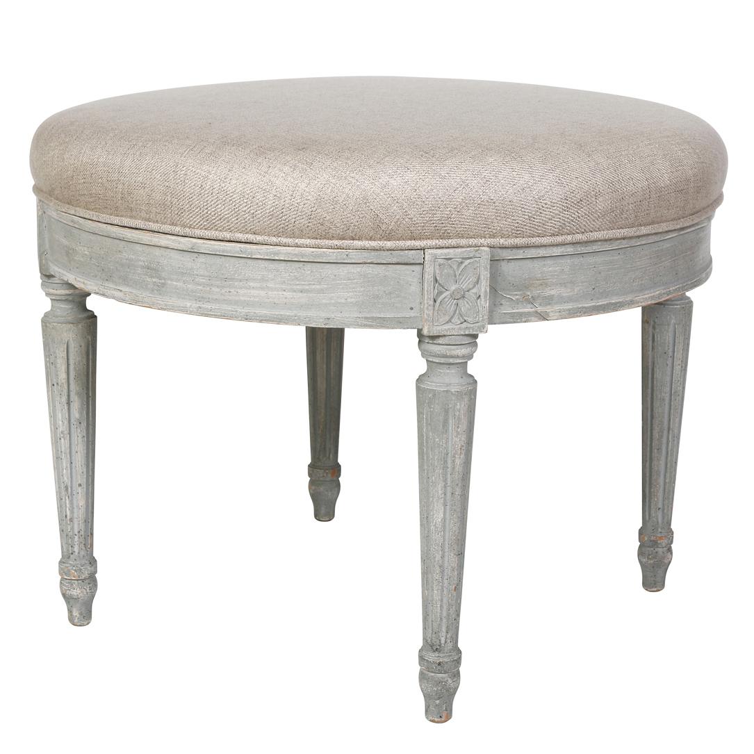 One of  two  in our inventory, this small round ottoman has a grayish white painted base.  Its carved apron and round, tapered legs are characteristic of the Louis XV style.  The top is upholstered in a neutral linen, making it ready to go into any