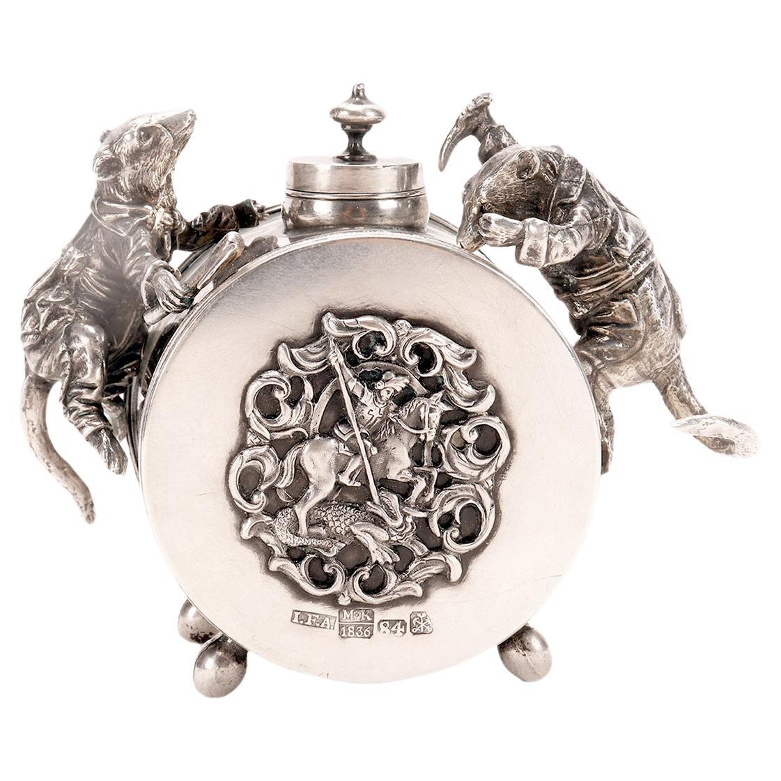 A Russian silver figured inkwell, jeweler: Johann Fredrik Akerblom, taster Mikhail Mikhailovich Karpinshií. Cylindrical inkwell raised on four feet with chiseled and knurled floral edges. The cylindrical top is hinged with an urn-shaped terminal;