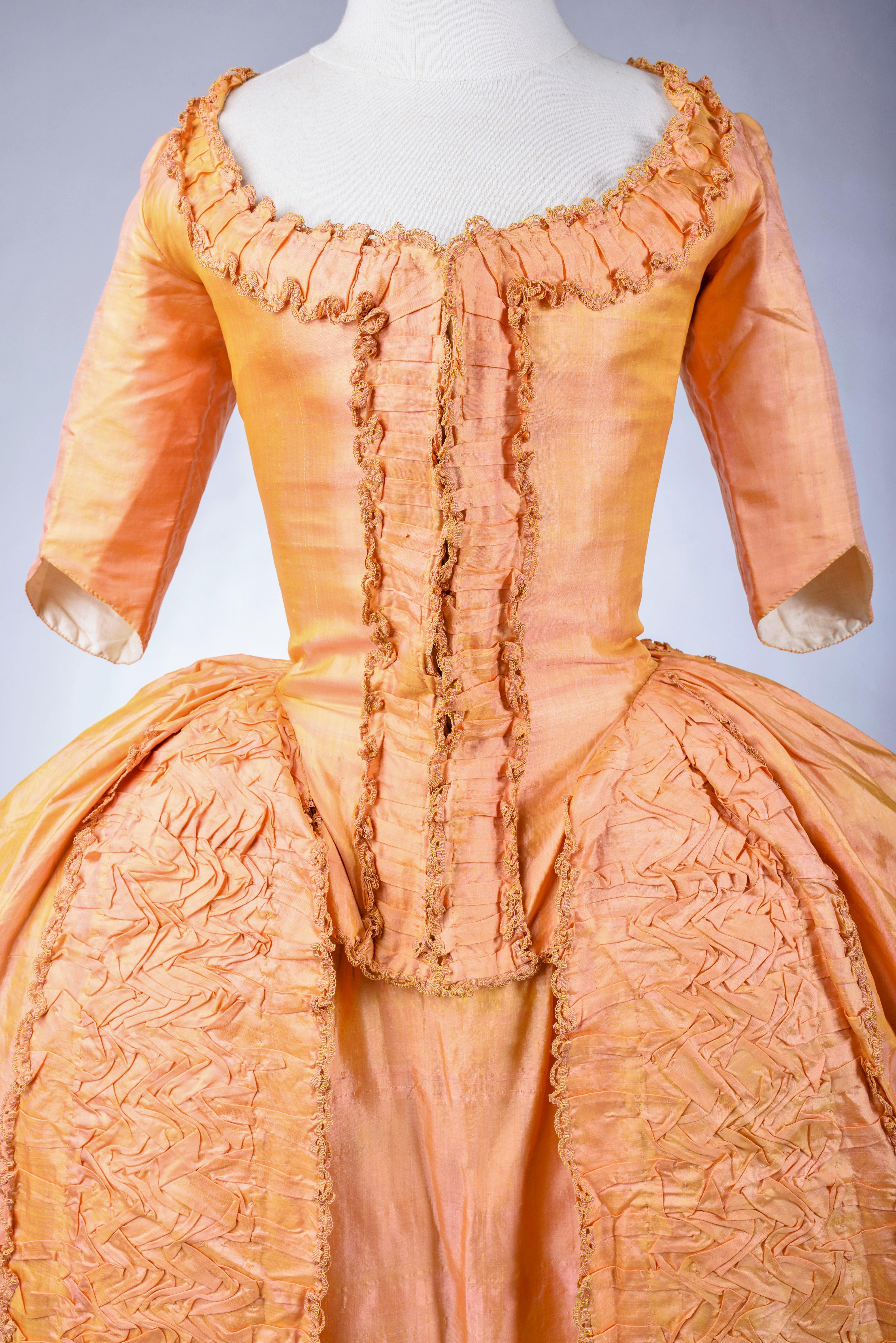 Circa 1785
France
A sack back gown, coat and skirt in salmon and yellow changing taffeta Florence silk. The gown has an à l’anglaise whalebone laced front bodice, three-quarter length sleeves and small à la Watteau pleats in the back ending in a