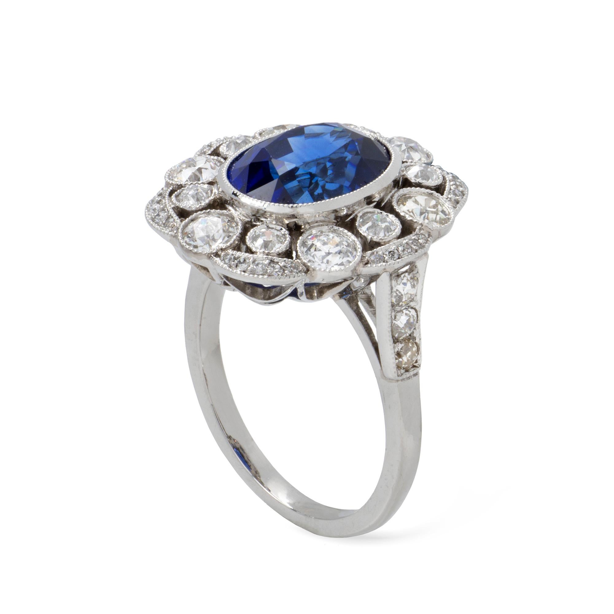 A sapphire and diamond cluster ring, the oval-shaped faceted sapphire weighing 5.17 carats, accompanied by GCS Report, stating to be of Sri Lankan origin, millegrain rubover-set to the centre of a cluster surround of brilliant-cut diamonds