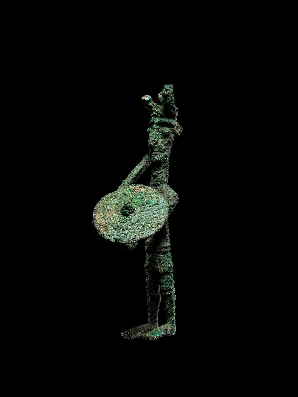 Una figura di guerriero in bronzo sardo eccezionalmente rara e importante. La figura altamente stilizzata è raffigurata in piedi e tiene nella mano destra una mazza appoggiata sulla spalla e nella sinistra uno scudo rotondo con borchia centrale. Il