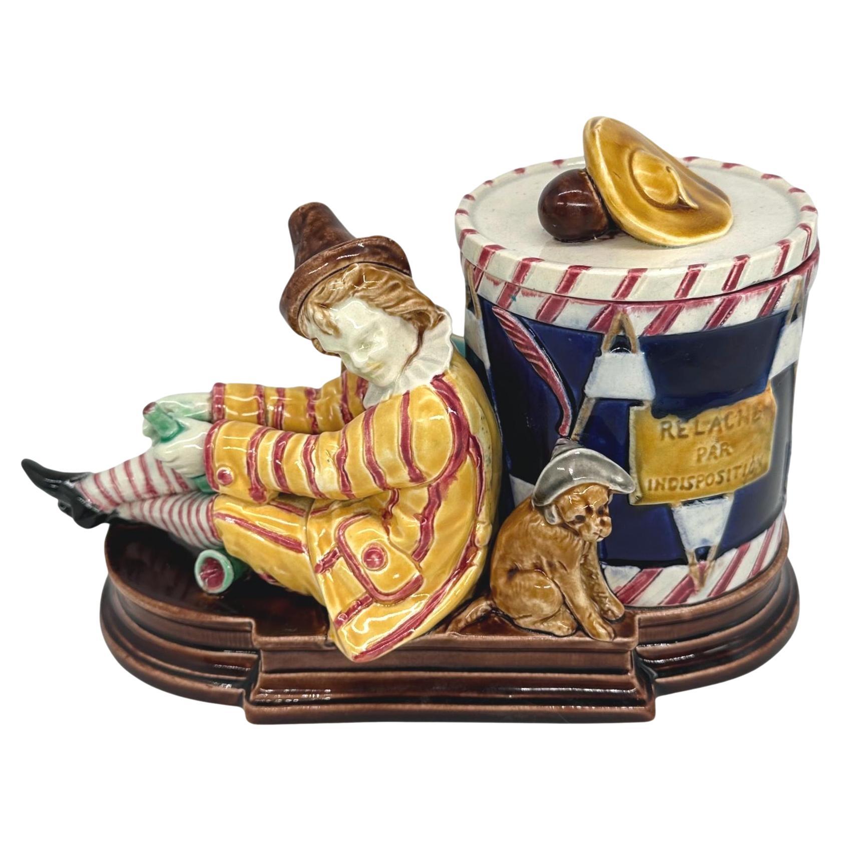 Sarreguemines Majolica Smoking Stand, molded and applied with an artiste de rue with his dog, and holding a wine bottle with an overturned wine glass below; the humidor formed as a cobalt glazed drum with a sign stating in French that the performer