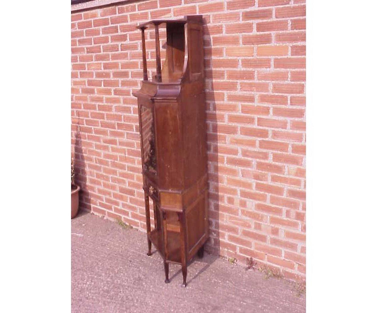 A good quality secessionist style walnut corner display cabinet, with upper and lower open display areas and a beveled glass door to the central display area with stylized brass escutcheon, a drawer below with a stylized brass handle, all stood on