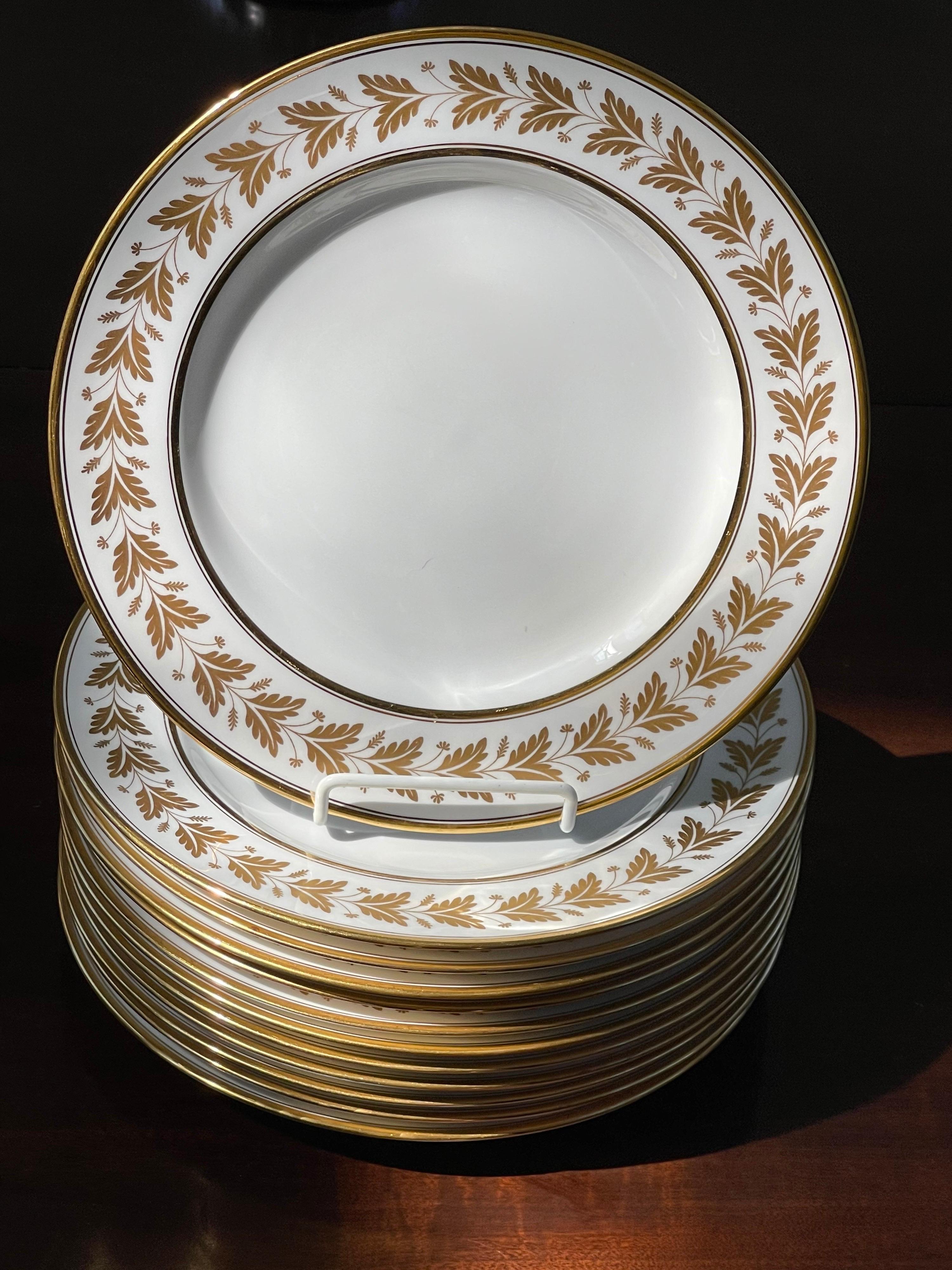 Un ensemble de 12 assiettes plates en porcelaine Spode Copeland avec une large bordure décorée de lauriers en or 24 carats sur fond blanc.