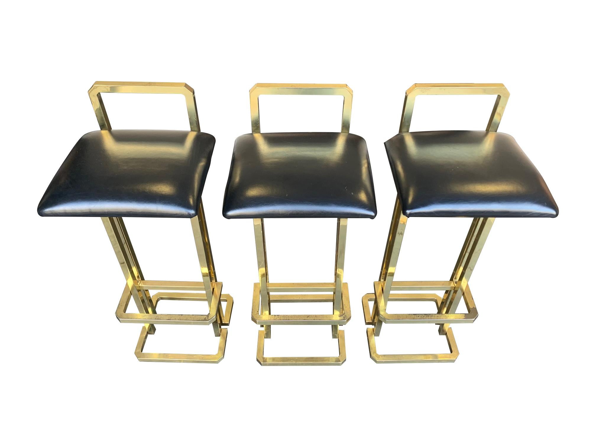 A set of 3 1970s Maison Jansen style gilt metal stools with black leather seat pads, each one has a back and foot rest.
