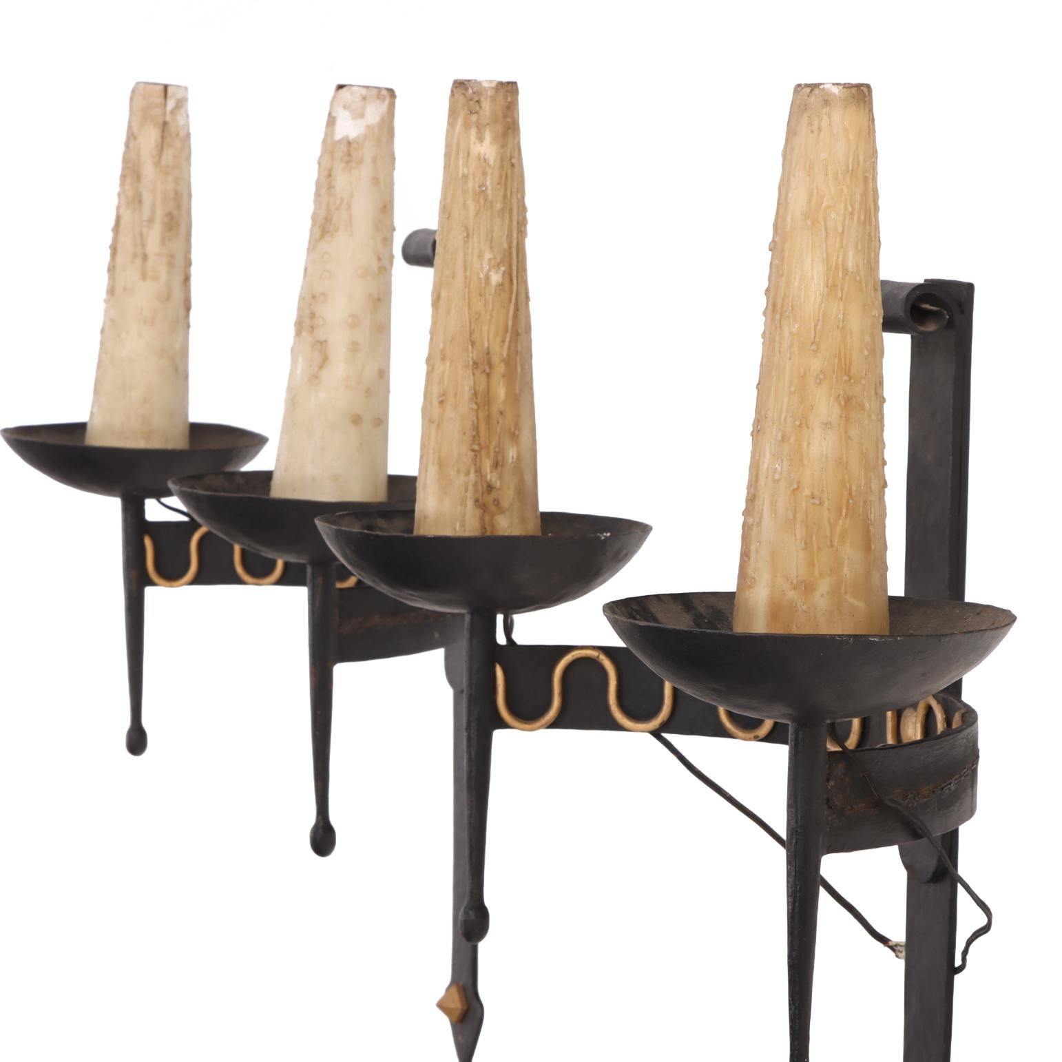 A striking set of four sconces. Each sconce features a pair of scrolling arms, each supporting a single electrified candle, flanking a central vertical support accented with brass details. The form tapers to a pointed terminus, giving the design a