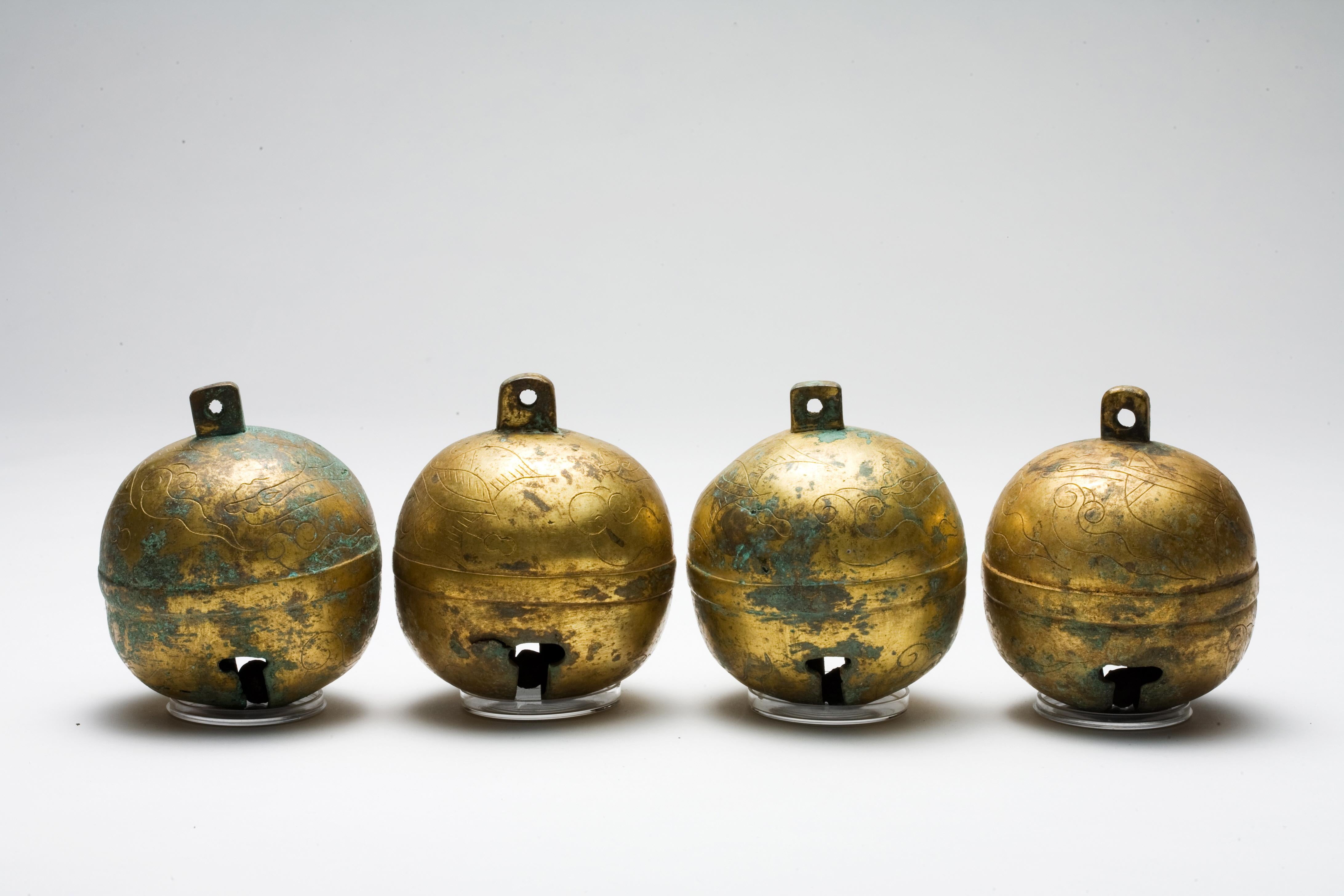 Un ensemble de quatre cloches en bronze doré, chacune coulée dans une forme presque sphérique avec une petite boucle au sommet pour la suspension et une fente étroite en dessous pour la résonance du son. Les surfaces présentent des traces de dorure