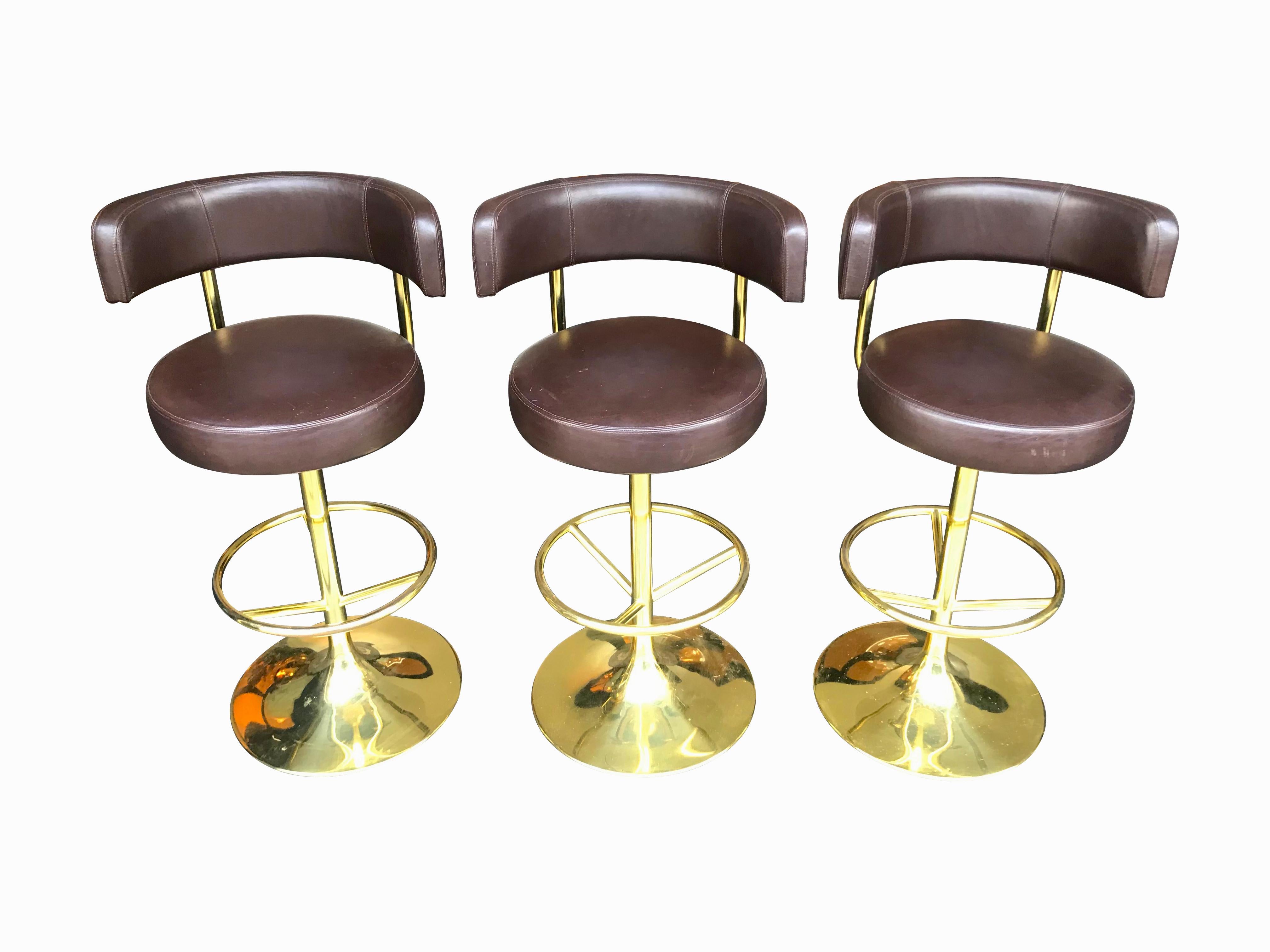A set of three original Börge Johansson “Jupiter” bar stools with tan brown leather seats and backrests on a revolving gilt metal base and frame. Each has a footrest and original 