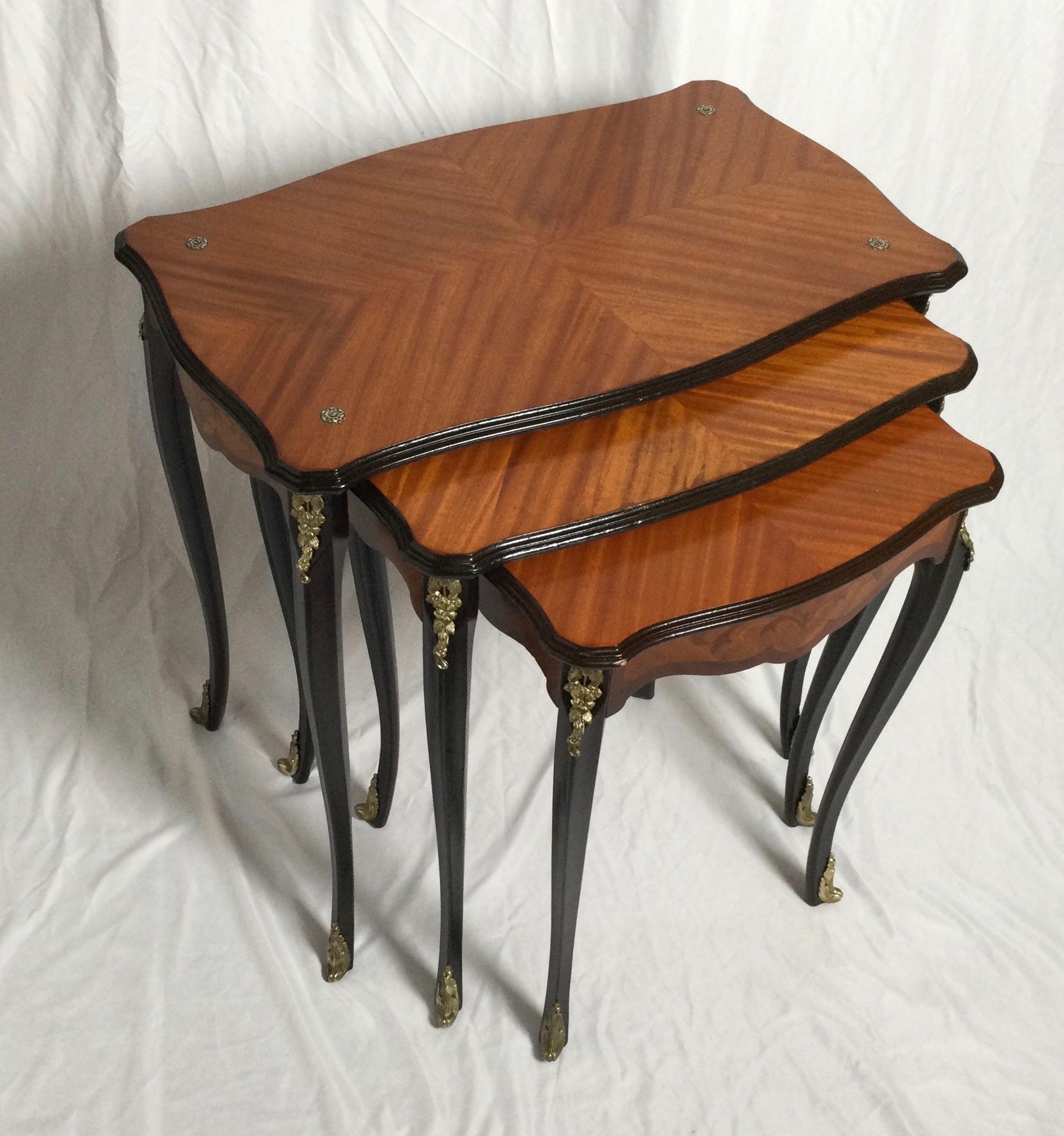 A graceful set of three book matched top nesting tables with dark mahogany legs and trim. There are cast burnished brass mounts at the top, and on the legs, Early to mid-20th century.