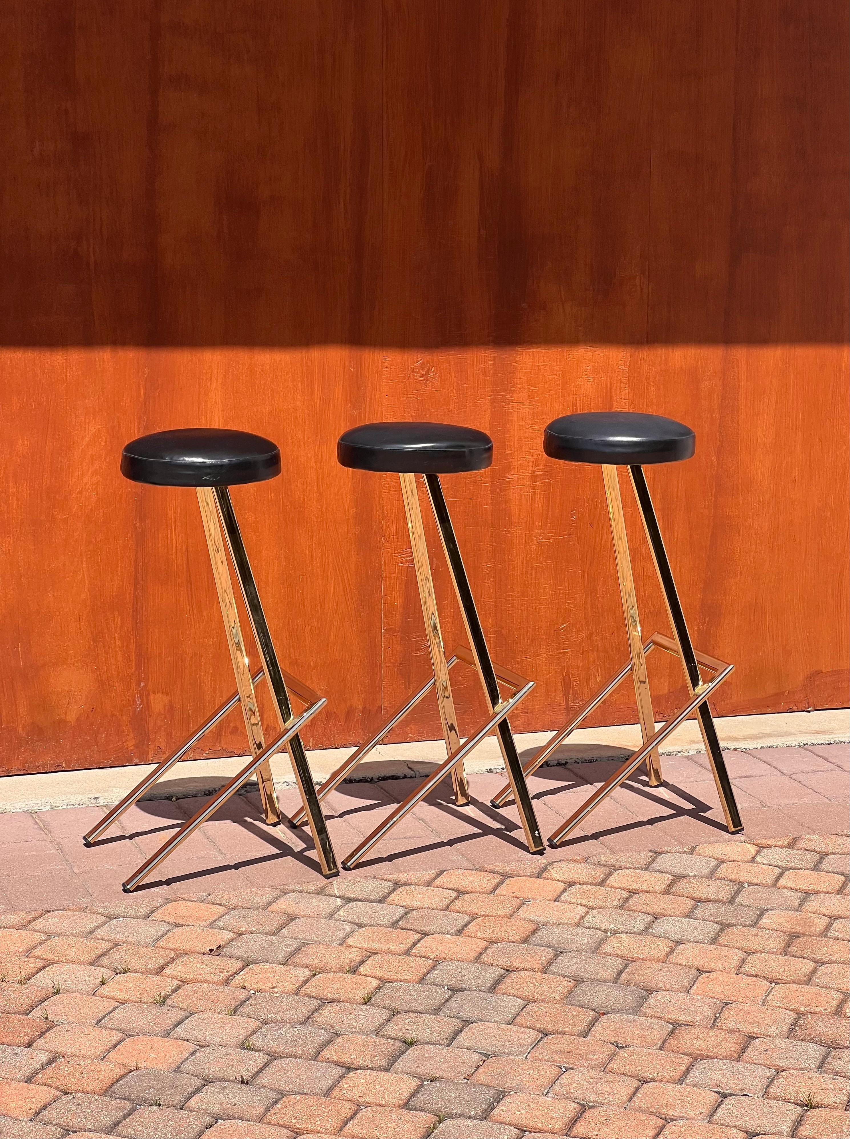 A set of three vintage postmodern bar or counter stool featuring gilt metal frames and faux leather seating, probably circa late 1970s. The statement geometry of these pieces will a touch of glam avant-garde to any interior. Each stool has intact