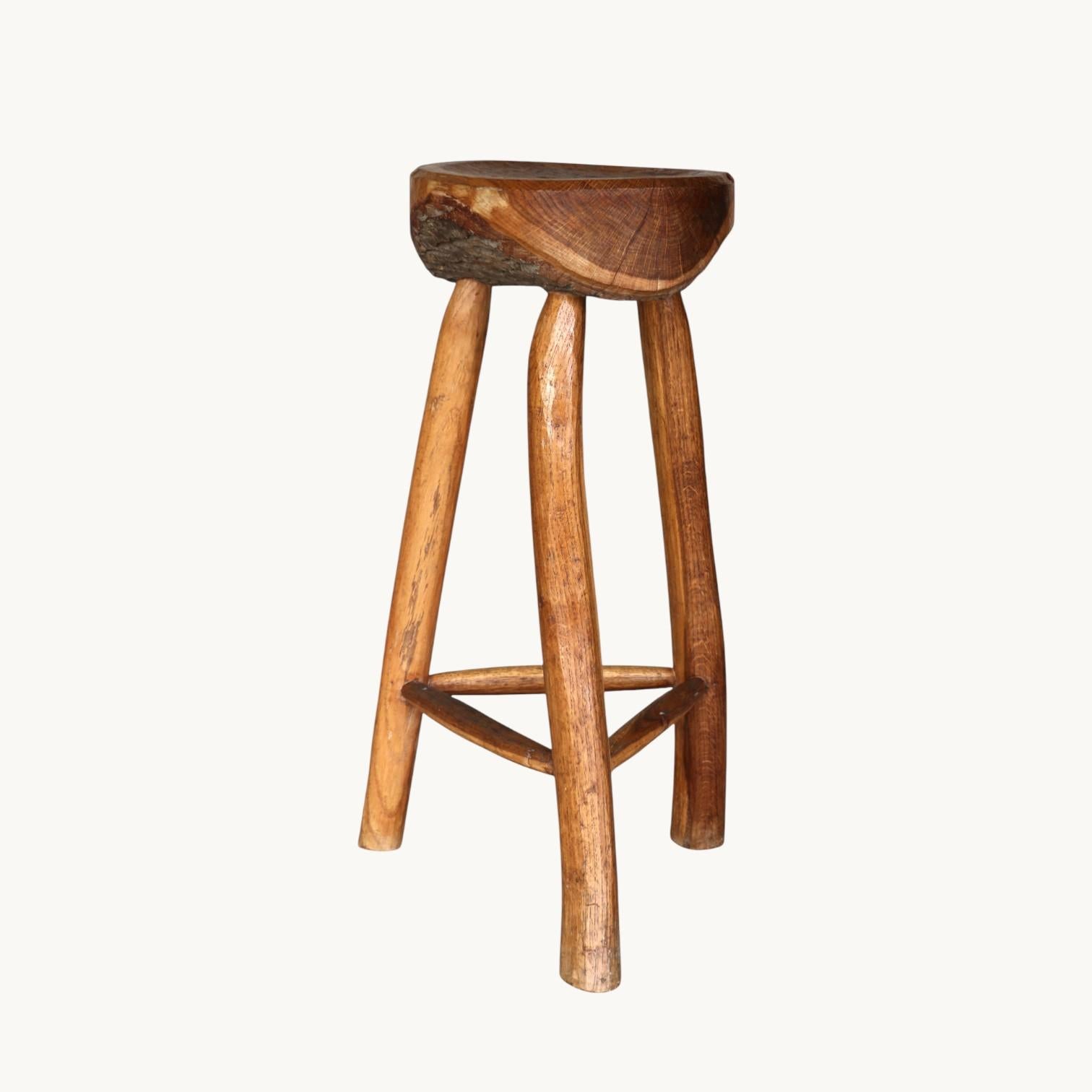A set of three high stools in raw wood. The seat is a thick, brutalist slice of tree trunk, the three legs are carved like branches. France, 60s.