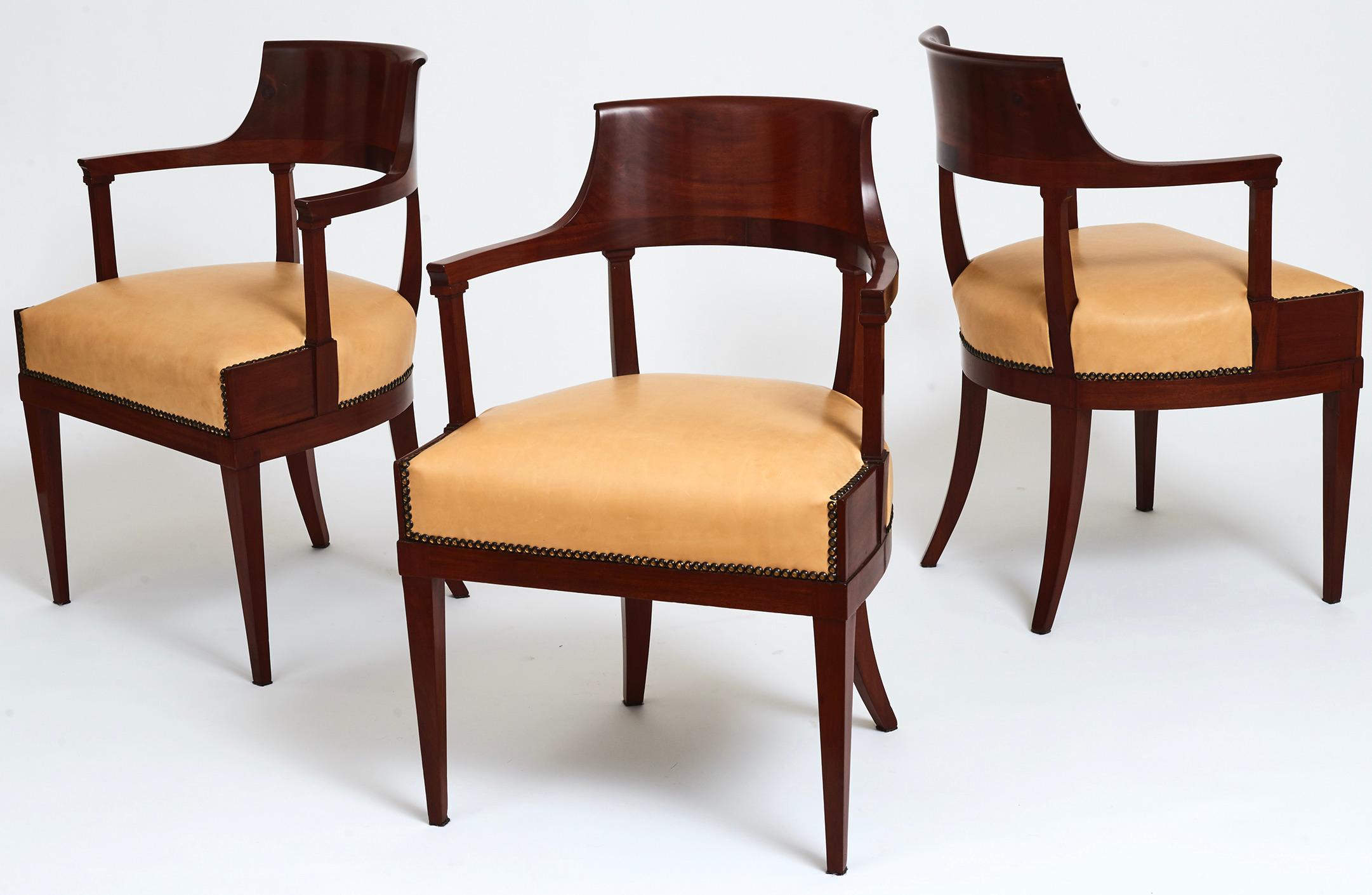A set of three Swedish mahogany neoclassical open armchairs, first quarter of the 19th century, each with a scrolled and curved backrest, straight cut back armrests above a wide frieze and upholstered seat, on square tapering legs. Restored and