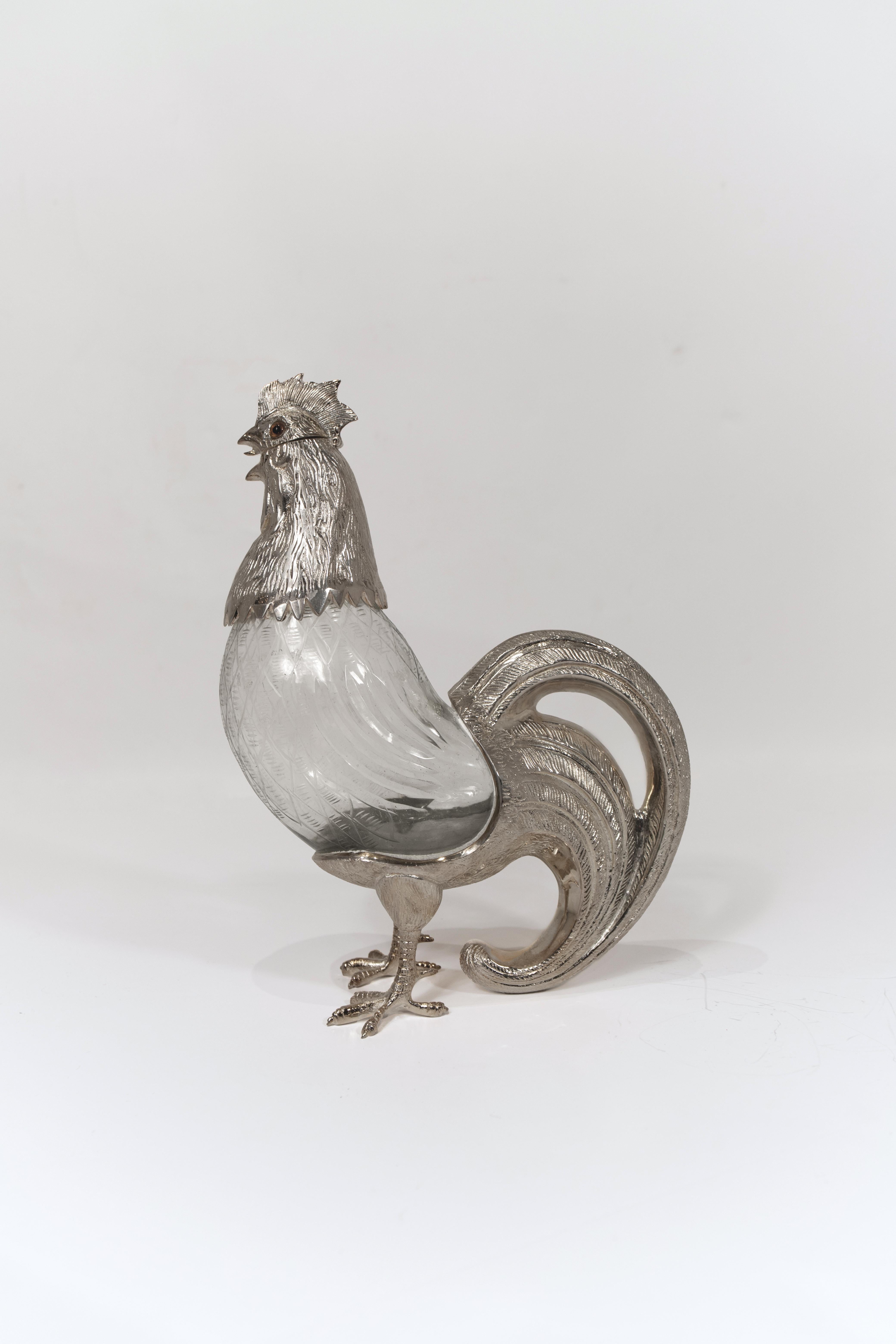 Carafe en métal argenté, conçue sous la forme d'un coq, dont la queue recourbée sert d'anse. Le bec verseur est astucieusement intégré dans la bouche du coq, recouverte d'un couvercle à charnière. Cette pièce remarquable allie fonctionnalité et art