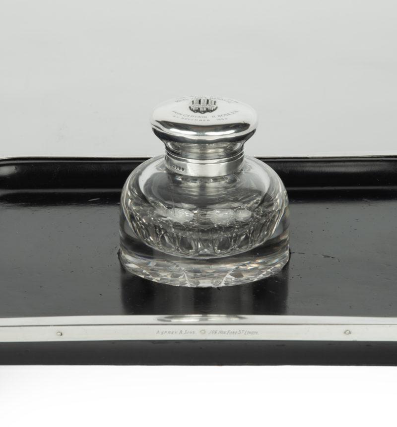 A silver and cut-glass desk set comprising a rectangular ebonised pen tray with silver mounts inscribed ‘Asprey & Sons, 166 New Bond Street London’ with a central cut glass inkwell.  The hinged silver lid of the inkwell is embossed with three