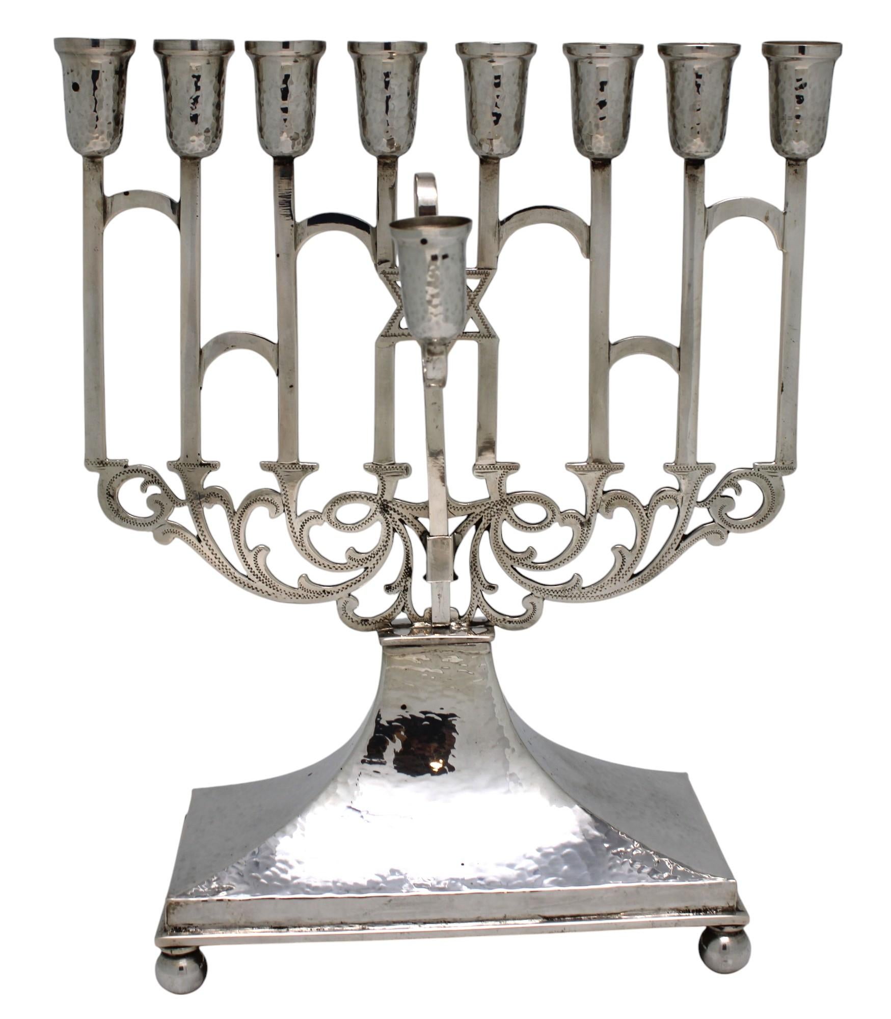 This elegant silver Hanukkah lamp, crafted in Austria circa 1925, showcases a refined blend of traditional and Art Deco influences. The menorah features a beautifully hand-hammered design on the lower base and oil cups, adding texture and depth to
