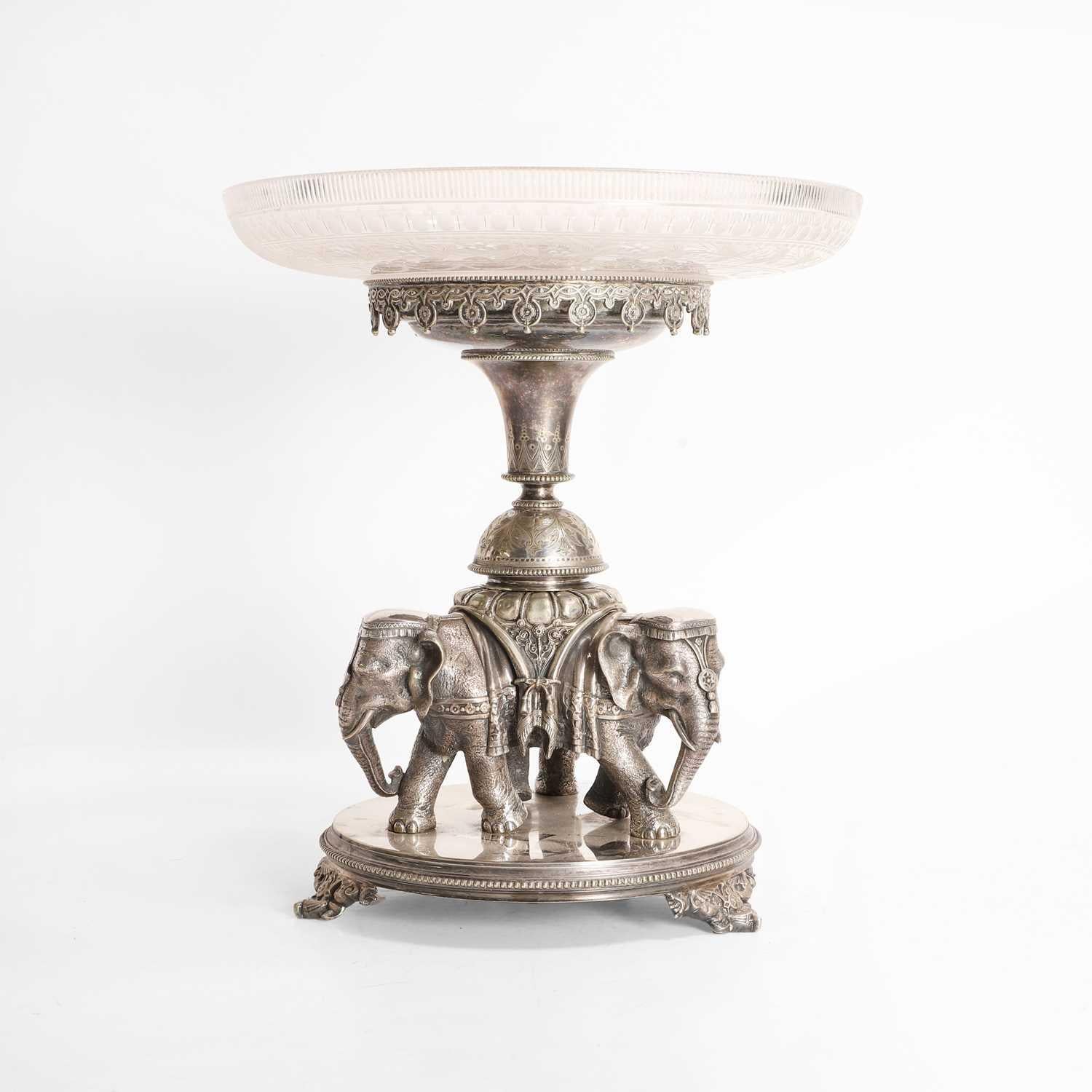 A silver-plated centrepiece, late 19th Century, cast with three conjoined elephants holding aloft a flared and engraved stand, with a cut and etched-glass dish, raised on a circular plinth with a beadwork edge and cast dragon feet,

