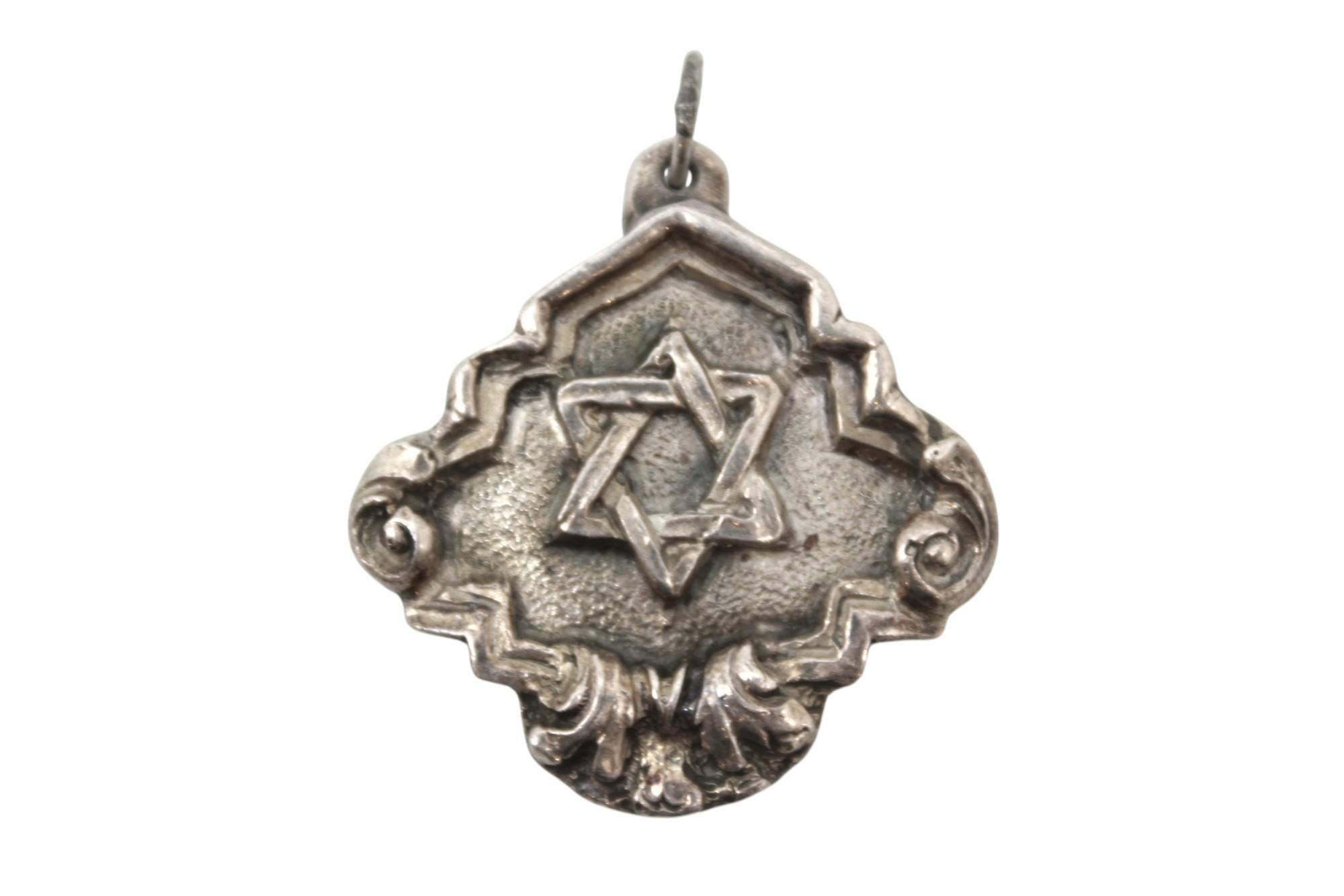 Un ciondolo in argento di Henryk Winograd, famoso maestro del repoussé, sorprendente e ricco di dettagli, realizzato con una forma a diamante arrotondata e che mostra l'abilità di Winograd sia nel design che nel simbolismo.

Al centro del ciondolo