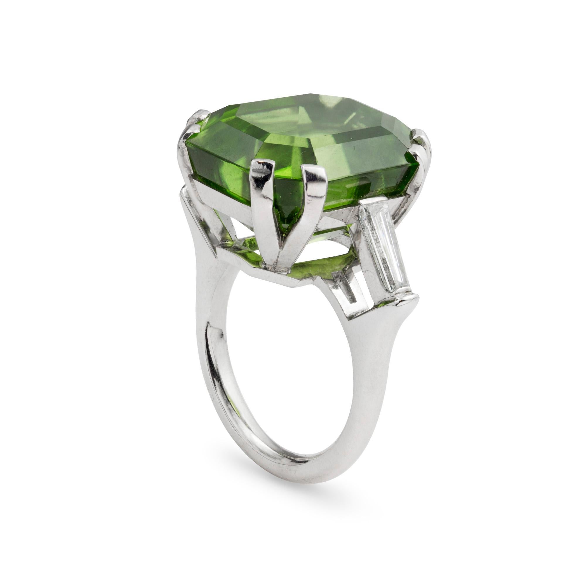 A single stone peridot ring, the mixed-cut octagonal shape peridot weighing 25.2 carats, eight yellow gold split claw set to a platinum mount, with tapered baguette-cut diamond set step down shoulders, the diamonds estimated to weigh 0.58 carats in