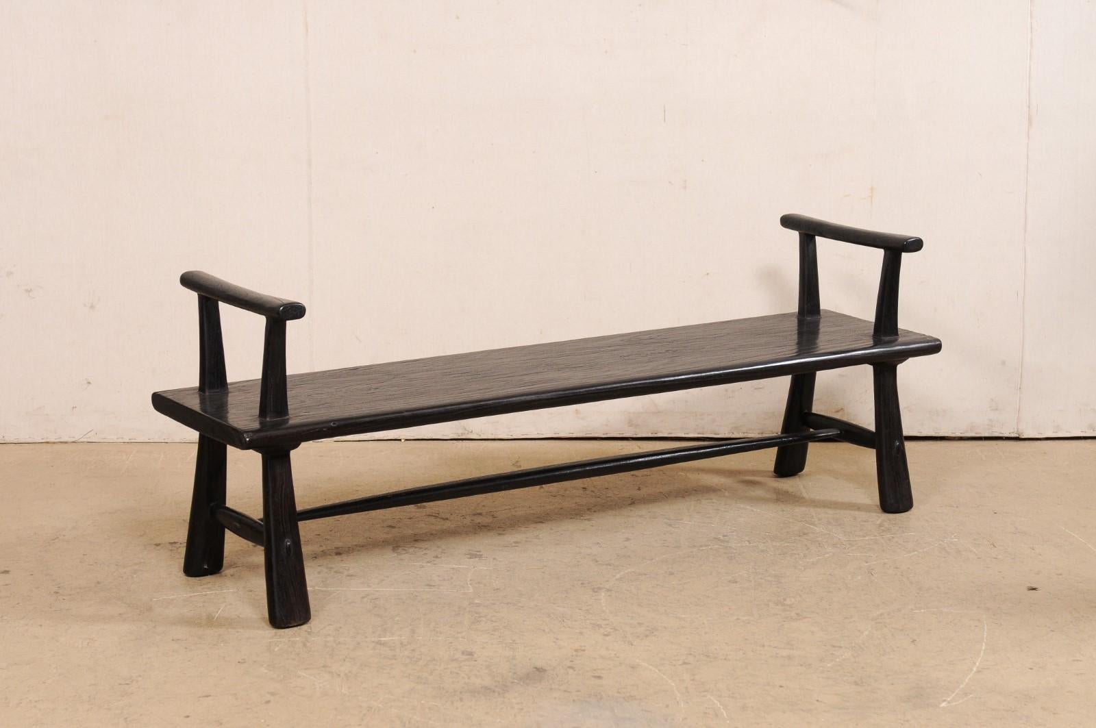 A sleek, artisan-made ebonized bench of teak wood. This hand-crafted wood bench from Indonesia has a rectangular-shaped seat, extending approximately 5.75 feet in length, flanked with raised arms at either far end, which are designed to appear as if