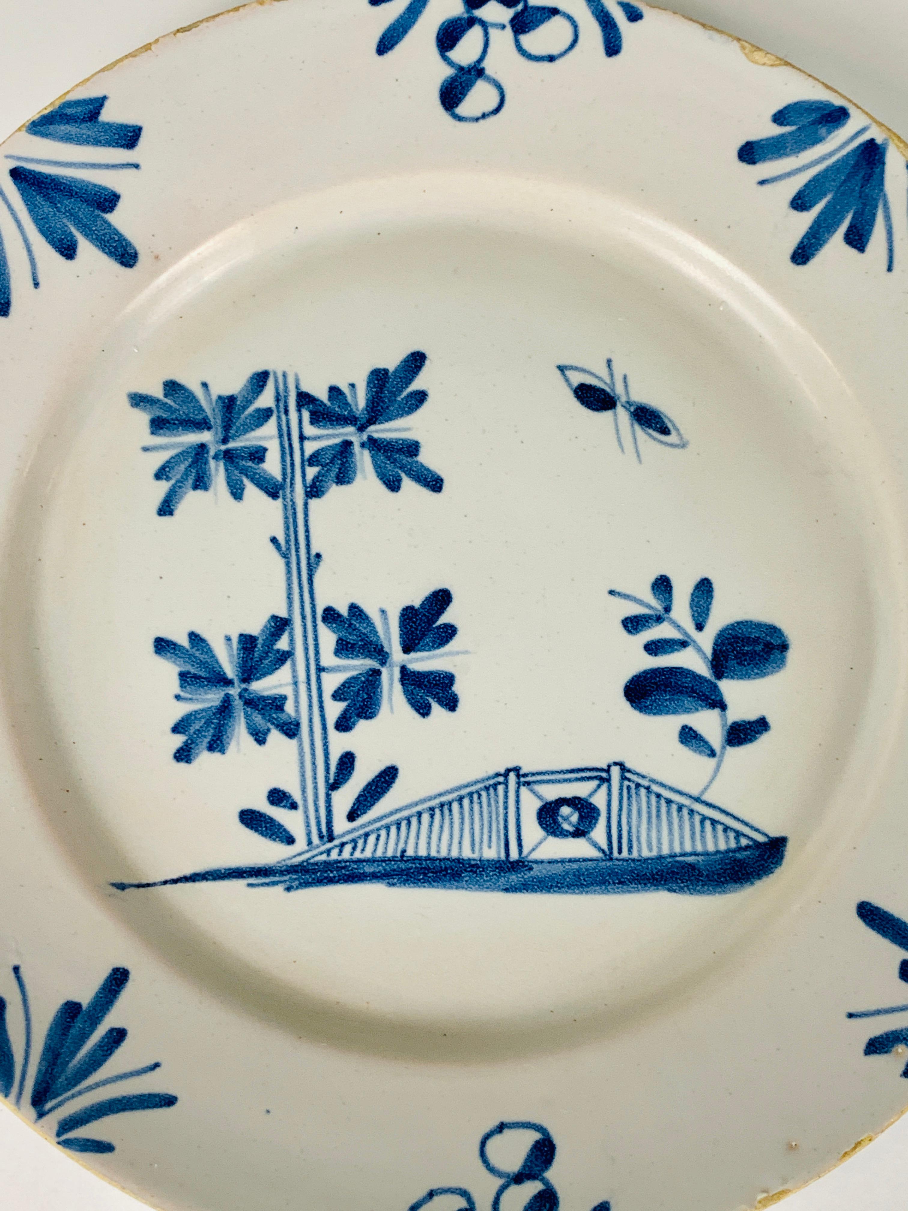 A small blue and white English Delft plate made circa 1740. 
The hand-painted decoration is naive, showing a garden fence, a leafy tree, and a butterfly. On the border are leaves and flowers.
The tin glaze is applied thinly, showing the underlying