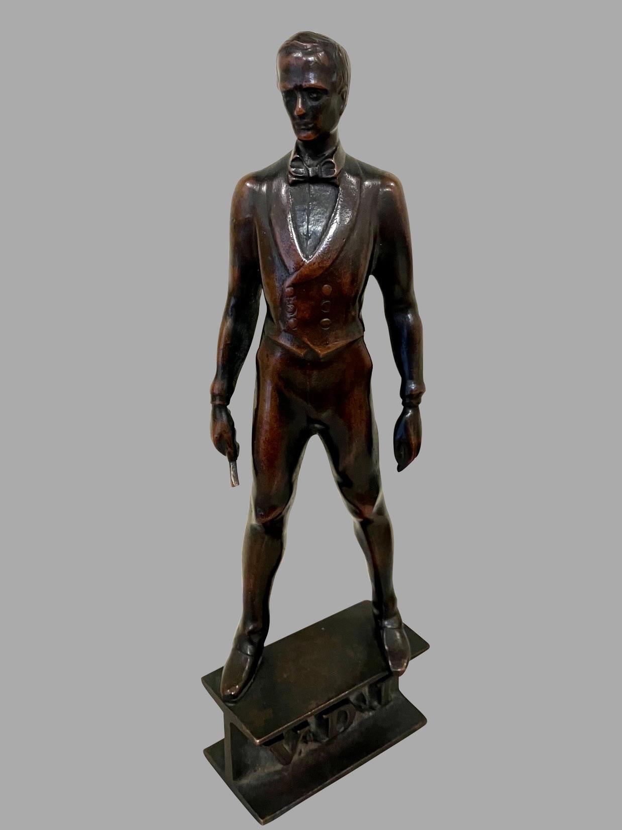 Un piccolo bronzo ben fuso di un gentiluomo formalmente vestito con un panciotto, che indossa un papillon e tiene quello che sembra essere un manganello nella mano destra. An He è in piedi su un basamento con le lettere incise A.V.D.I. Probabilmente