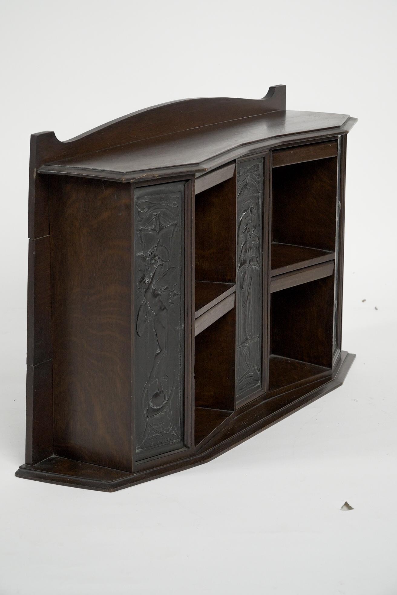An Arts and Crafts desktop oak bookcase with a shaped upper back and hinged dust flaps to the tops, with three bronze plaques. 
The left plaque depicts an angel with a quote at her feet: Born to speak all mirth. 
The centre plaque with an angel, and