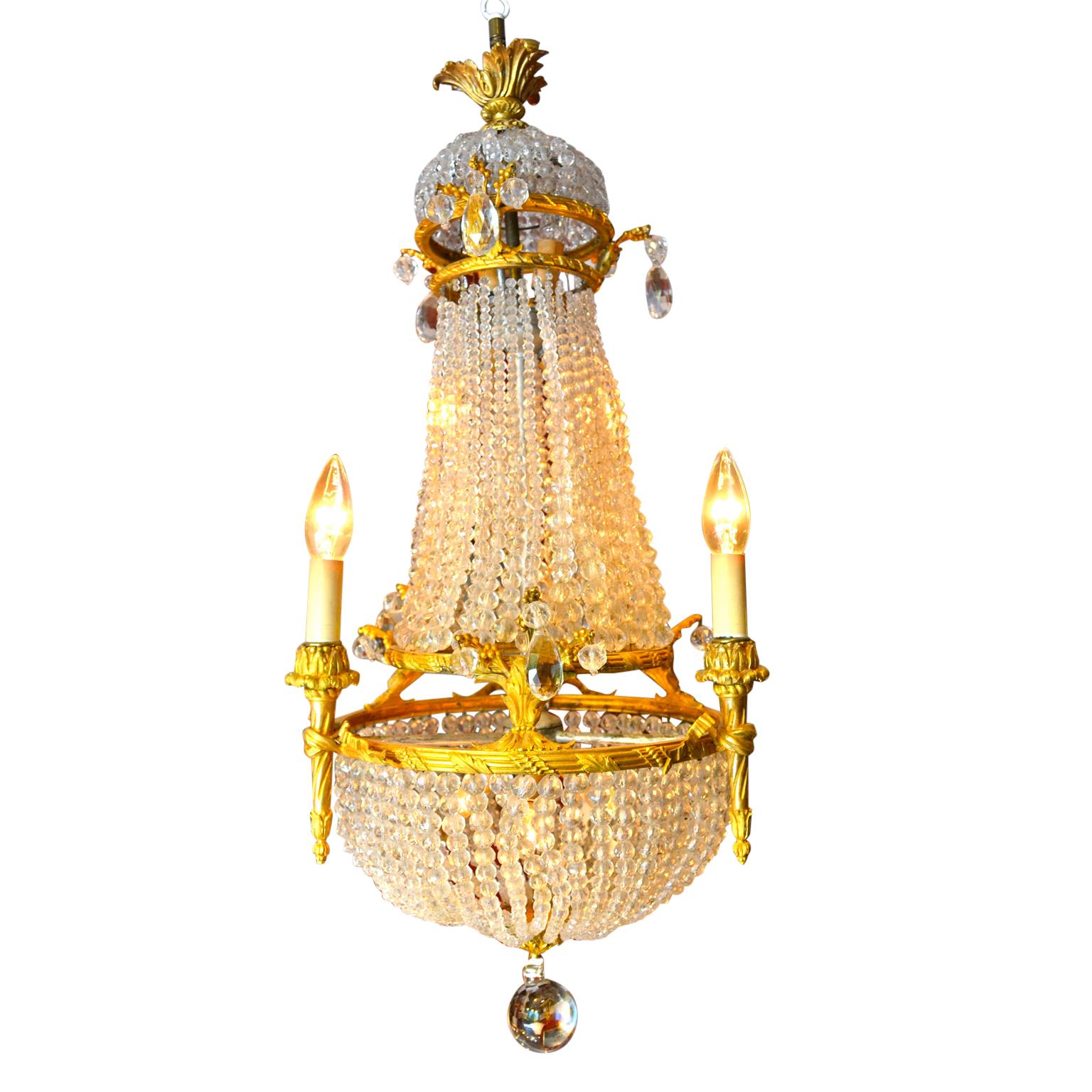 A small scale Empire style basket chandelier with lighting to the top, middle, and bottom sections. Each section is framed in gilded bronze supporting chains of cut crystal beads. There are three candle arms around the middle corona each supported