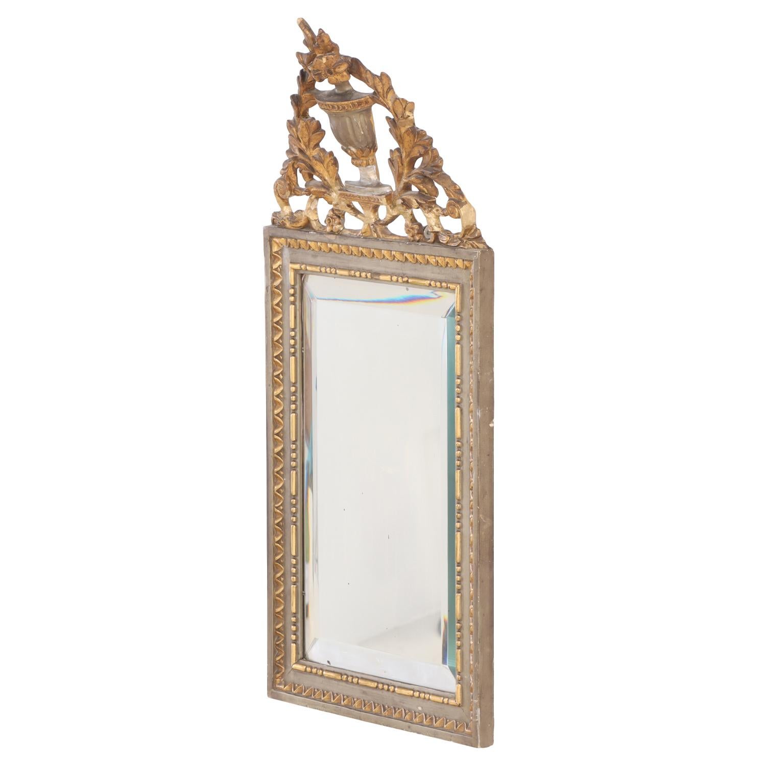 This charming small mirror features an intricately carved wooden frame, elegantly painted and accented with gilt detailing. The ornate crest with central urn of flowers surrounded by foliate embellishments exemplifies the delicate craftsmanship of