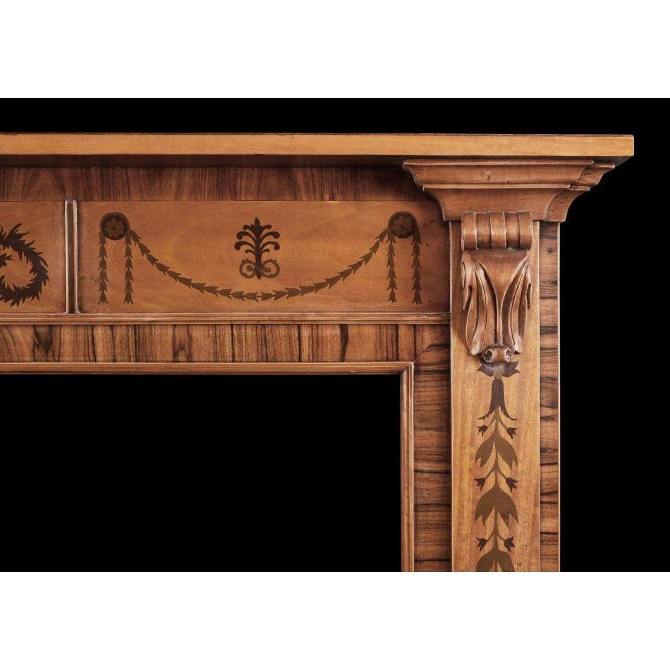 A small scale timber fireplace in the Regency style. The jambs inlaid with leaf work throughout surmounted y carved bracket. The frieze with inlaid drapery, rosette paterae and Anthemion leaves, with plain shelf above. One of an identical pair with