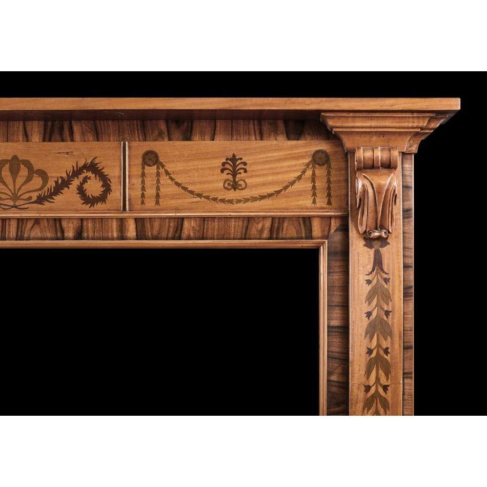 A small scale timber fireplace in the Regency style. The jambs inlaid with leaf work throughout surmounted y carved bracket. The frieze with inlaid drapery, rosette paterae and Anthemion leaves, with plain shelf above. One of an identical pair with