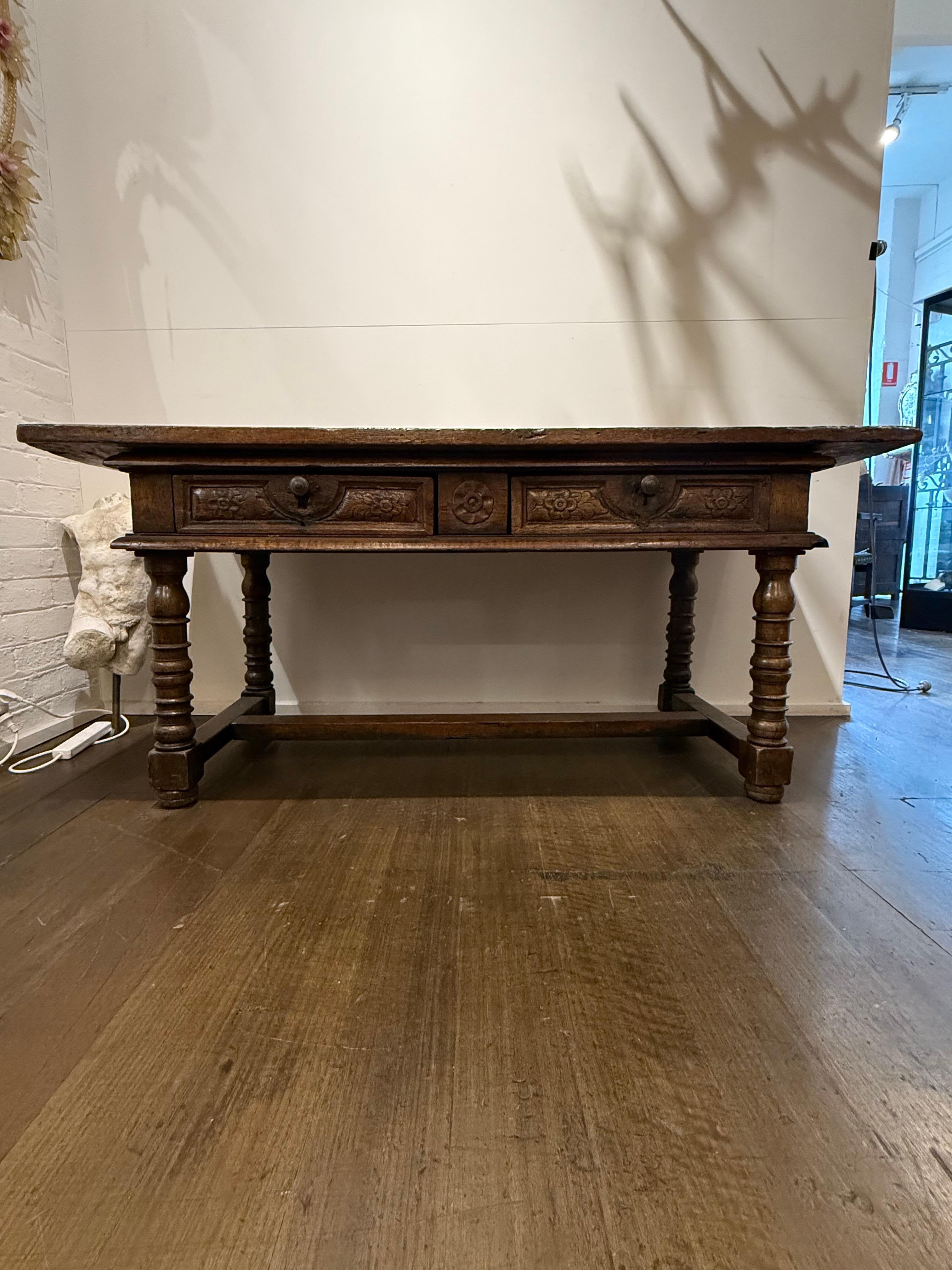 A Spanish Baroque Style Walnut Side Table, 18th Century

Description:
A substantial single plank top over a moulded frieze comprising two panelled drawers, each with a pair of carved floral motifs and centred by a brass knopped handle above an