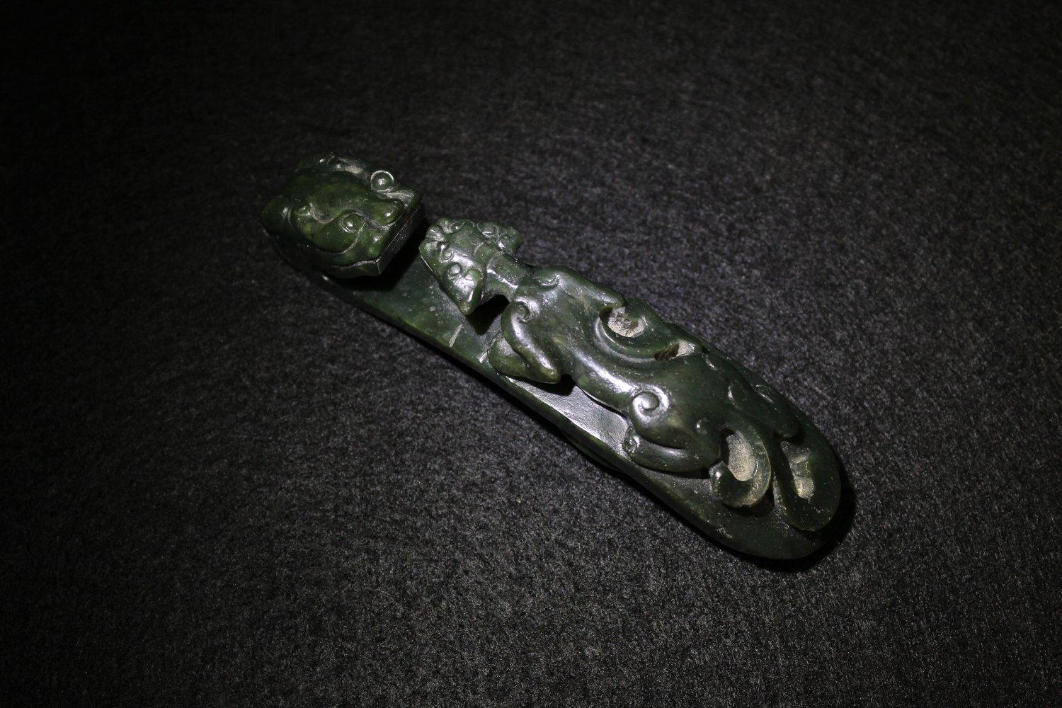 This spinach-green jade belt buckle is surmounted by a a prowling chilong with a goatee. Below the chilong is a bowed platform which curls up to become a large chilong head with ears, curved horns, and eyes with exaggerated eyebrows. 

Apart from a